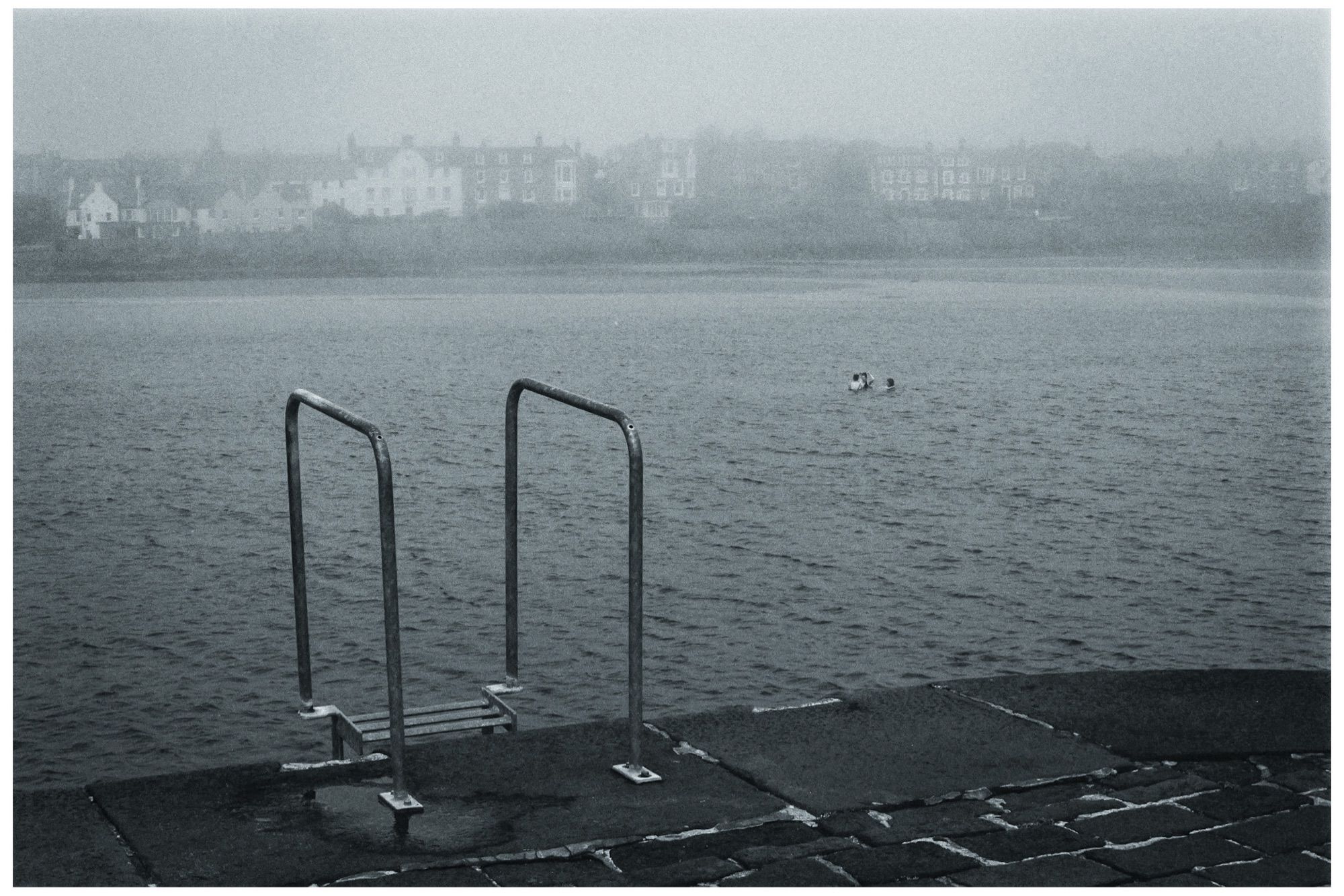 Ladder railings at Elie Harbour. Several people swim in the sea in the mid distance. A sea mist makes the houses in the background hazy. Film photography.