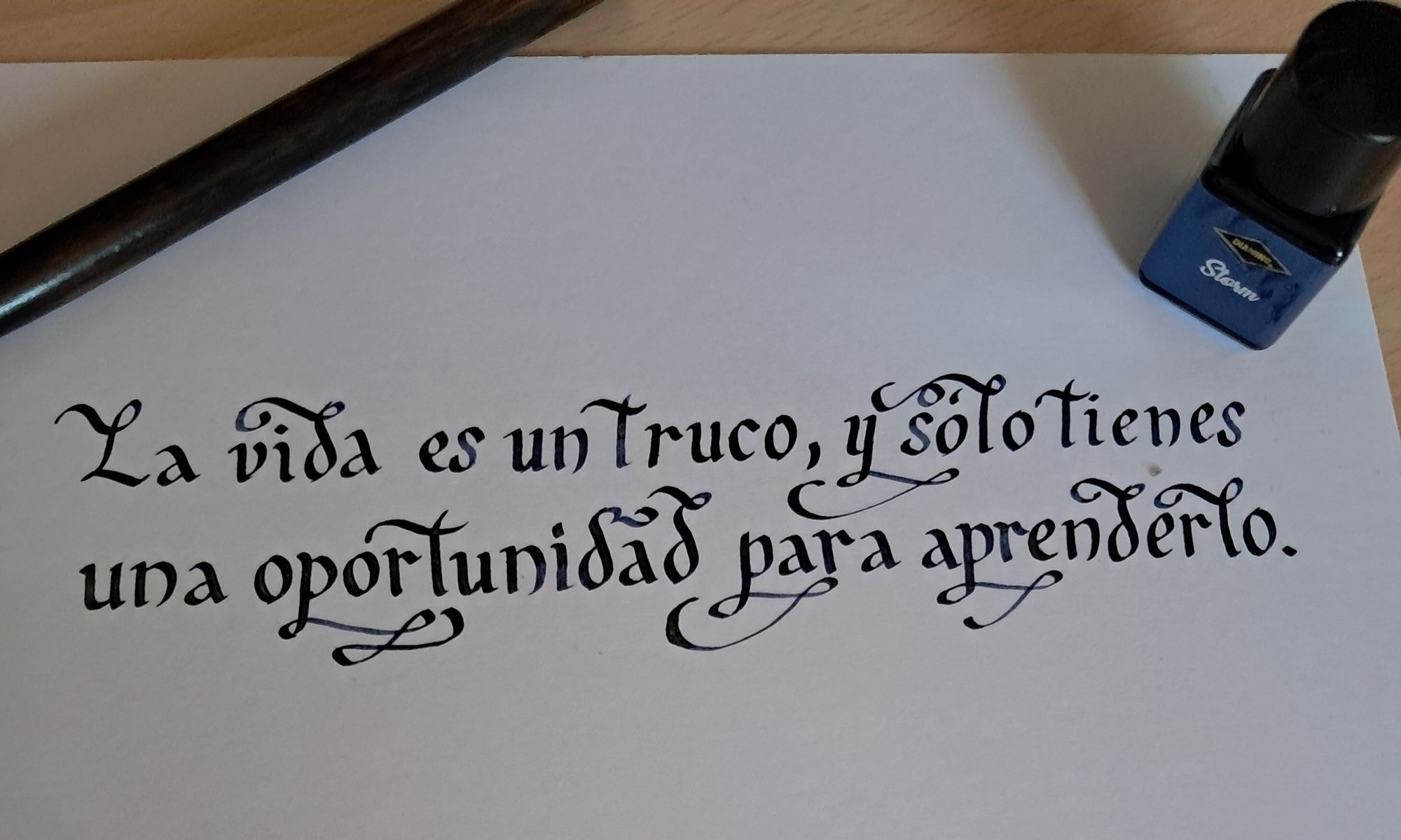 La frase "La vida es un truco, y sólo tienes una oportunidad para aprenderlo" escrita en tinta azul marino en caligrafía con florituras. A su lado el palillero de la pluma y el tintero (etiquetado como "Storm") usados para escribirla