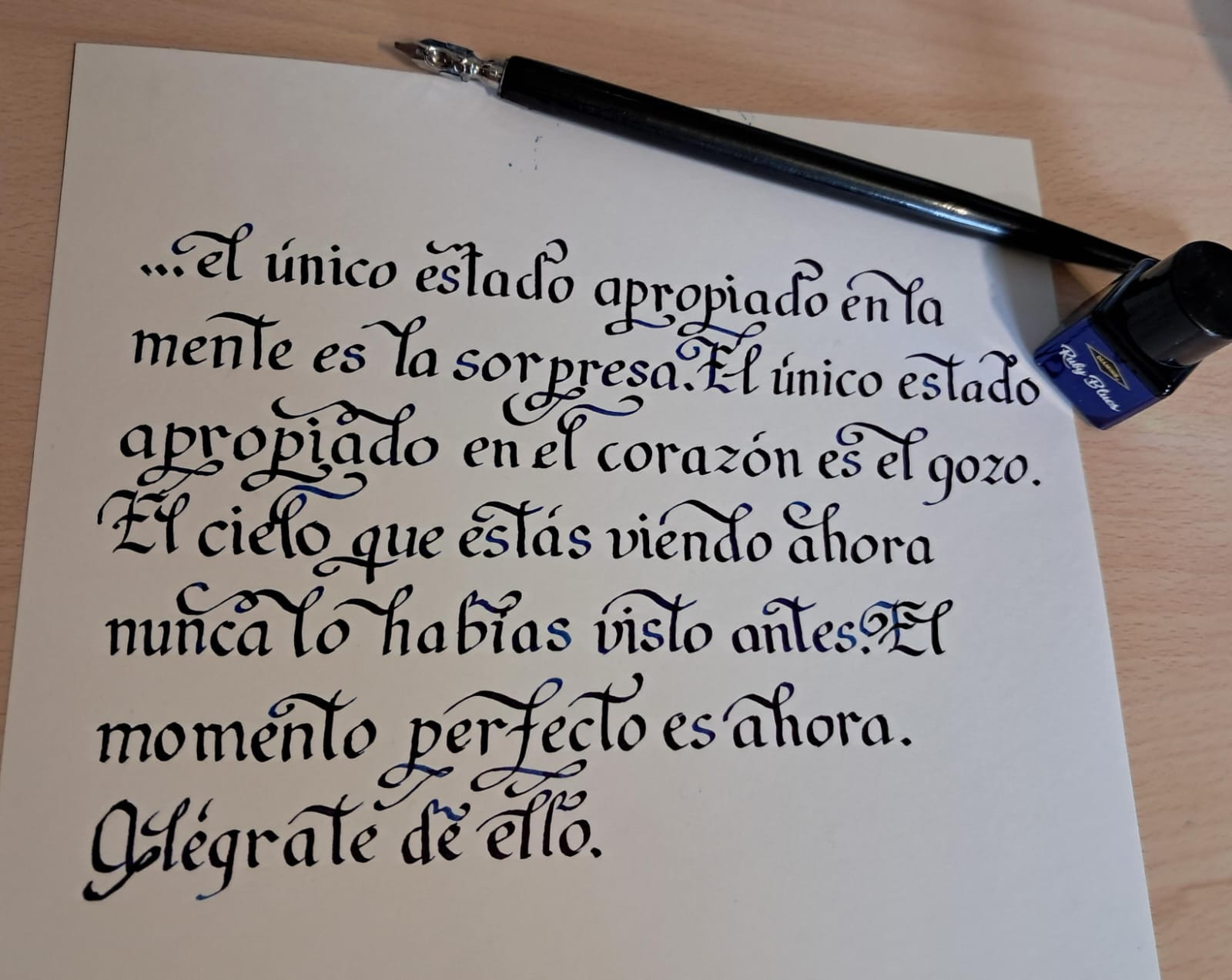 El texto "... el único estado apropiado en la mente es la sorpresa. EL único estado apropiado en el corazón es el gozo. El cielo que estás viendo ahora nunca lo habías visto antes. El momento perfecto es ahora. Alégrate de ello." escrito con tinta azul en caligrafía entrelazada. A la derecha, el plumín usado para escribirlo y un tintero pequeño etiquetado como Ruby Blues.