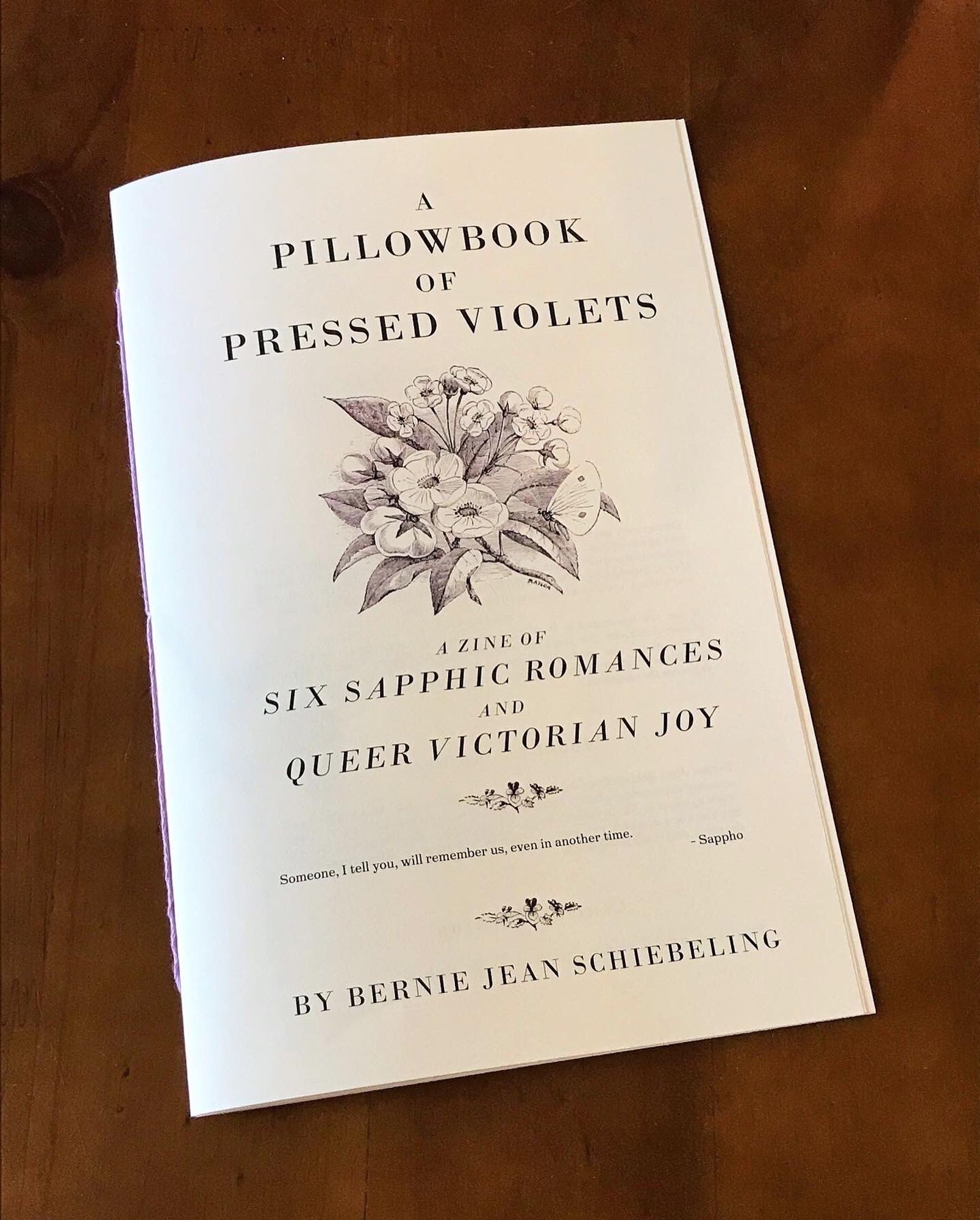 A half-page zine bound with lilac thread on a wood backdrop. The cover is styled to look like a Victorian pamphlet with a floral illustration, and the title reads “A Pillowbook of Pressed Violets: A Zine of Six Sapphic Romances and Queer Victorian Joy.” In smaller print, a quote from Sappho reads “Someone, I tell you, will remember us, even in another time.”