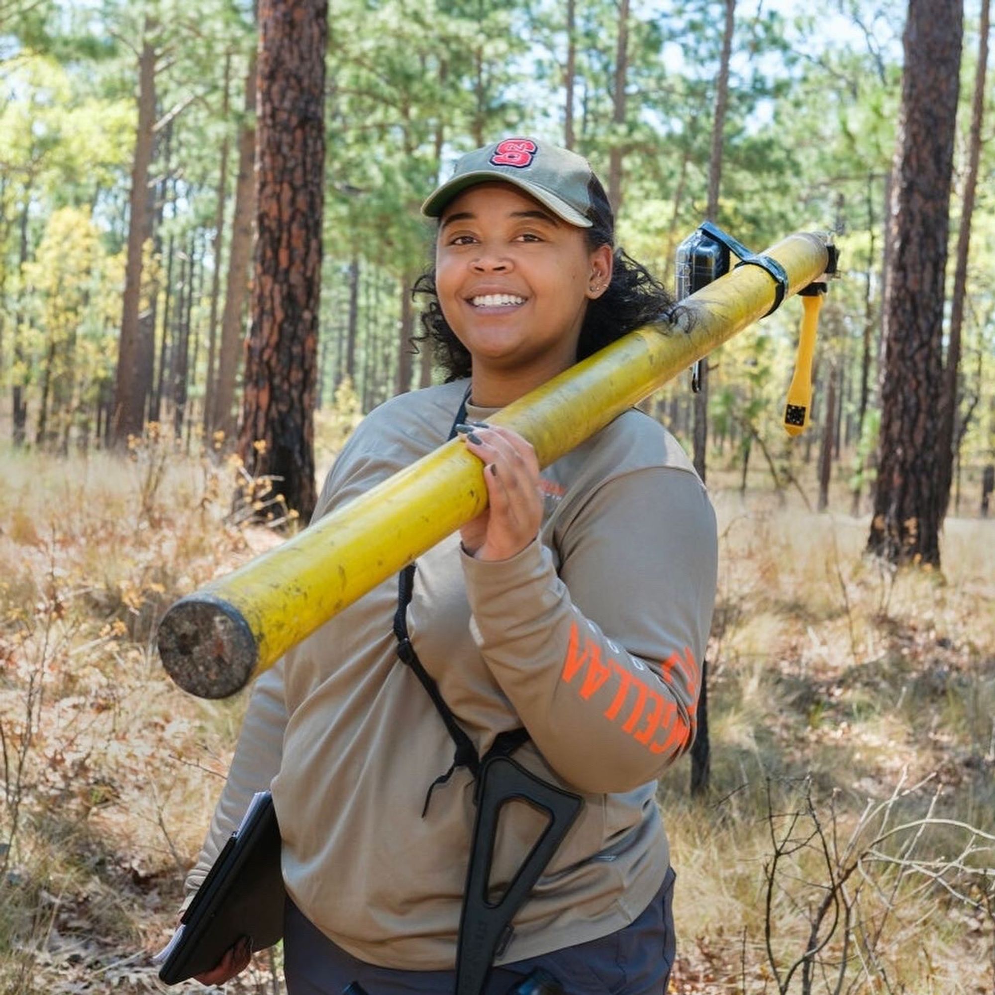 Lauren in a Longleaf pine ecosystem smiling while holding a cavity inspection pole, clipboard, and spotting scope.