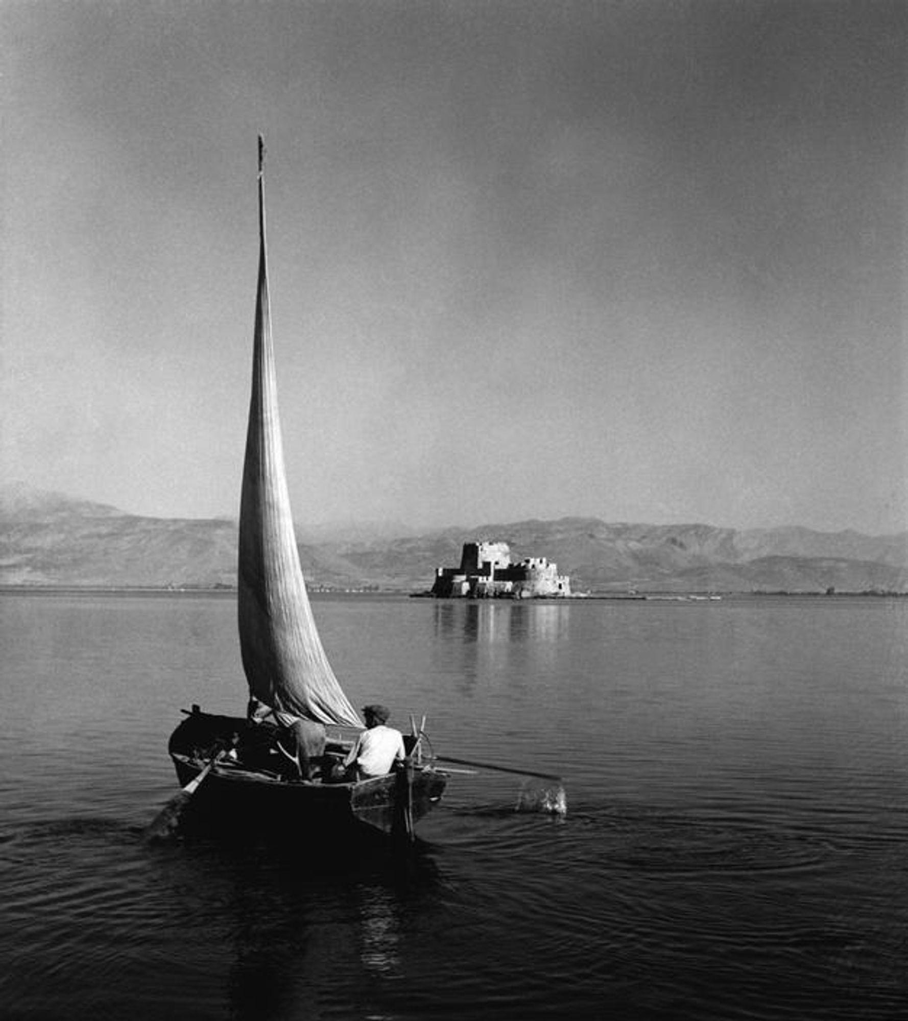 photo
a small boat, sail unfurled on a calm sea. a large castle like build in the middle of the frame and mountain ranges i the distance