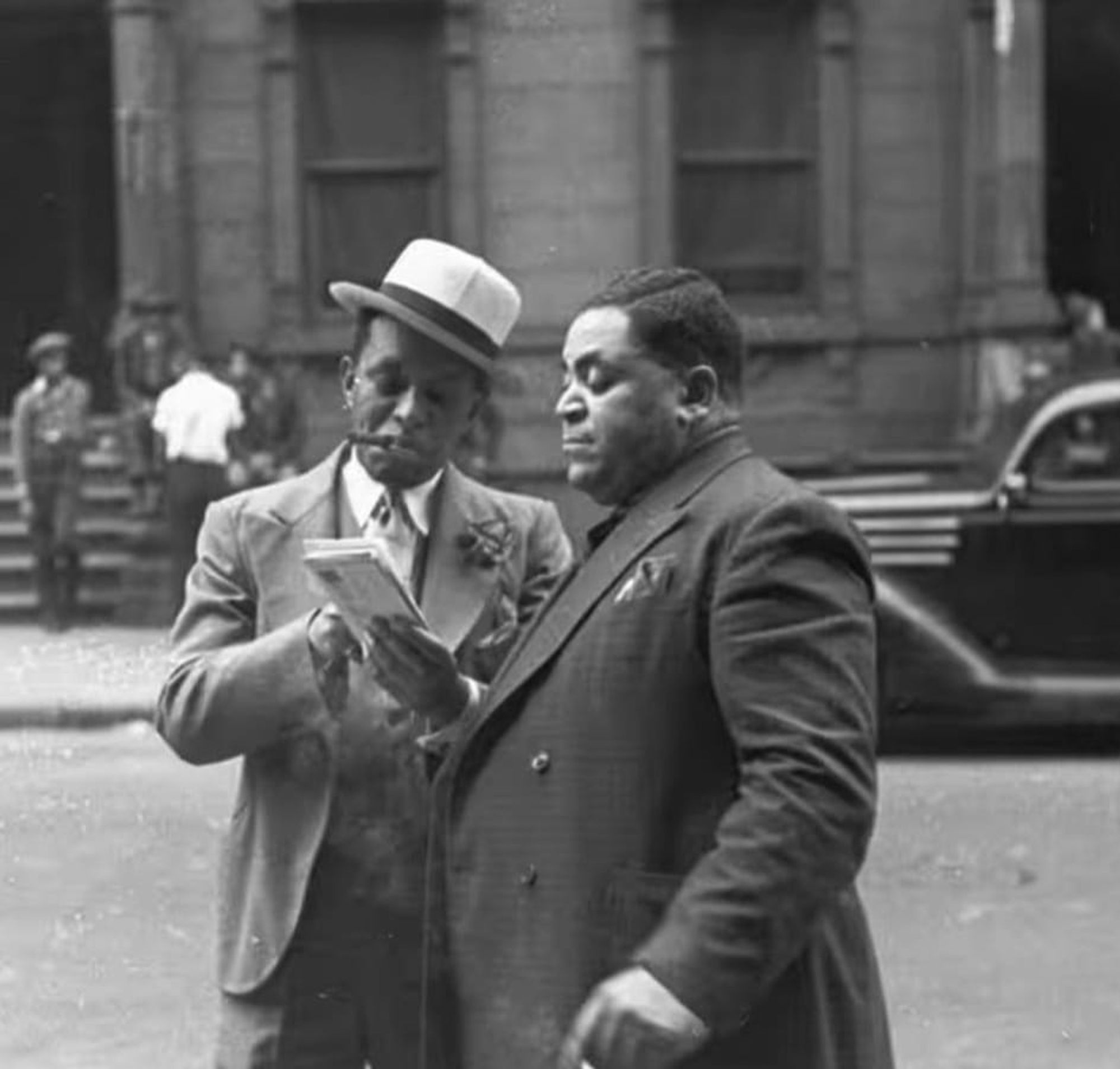 photo
street scene, two men looking at a piece of paper. a small group of people behind them and part of a car are also visible