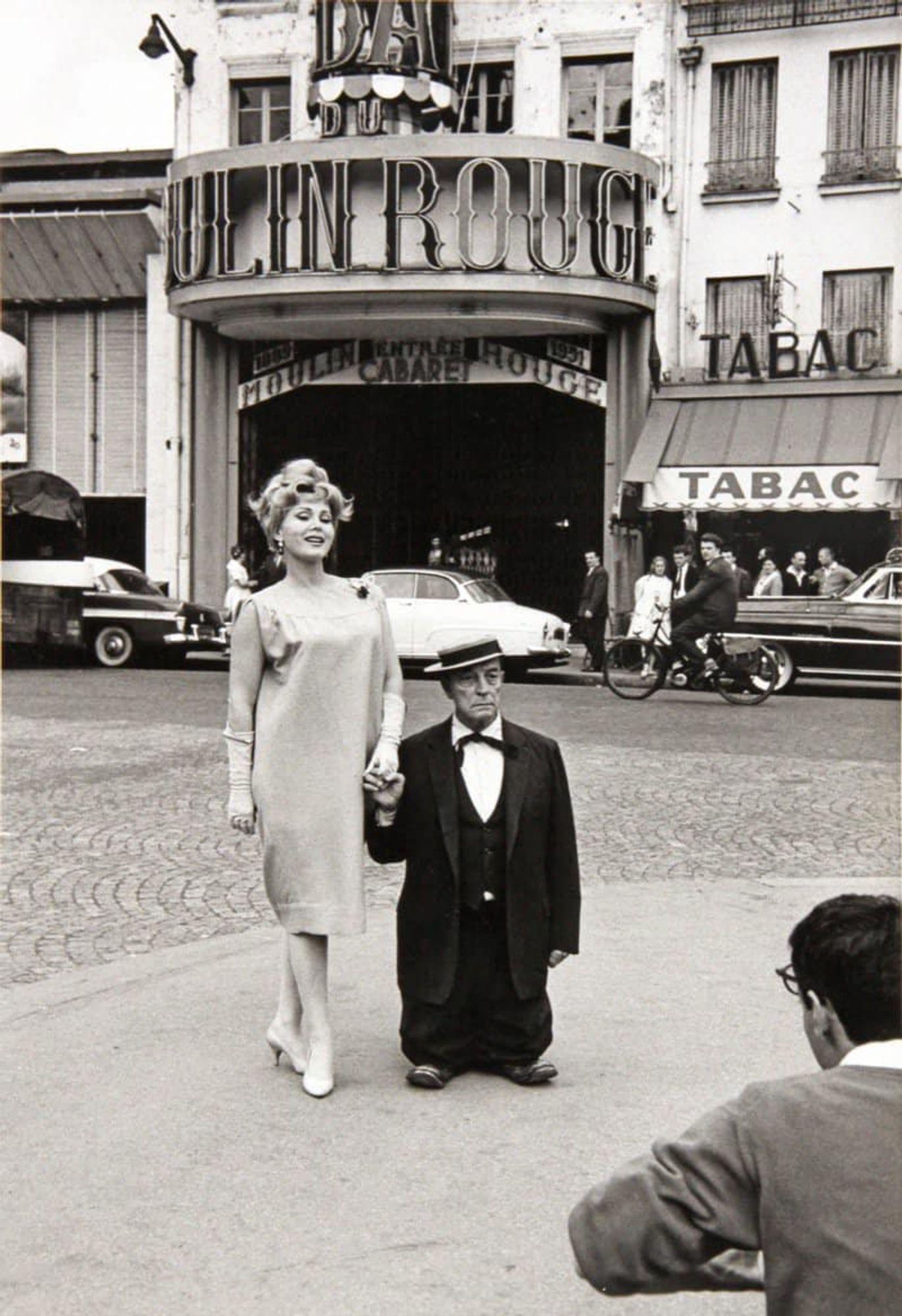 photo
a man and a woman outside the moulin rouge theater 
He is kneeling on his shoes, giving the impression he is very small compared to the woman