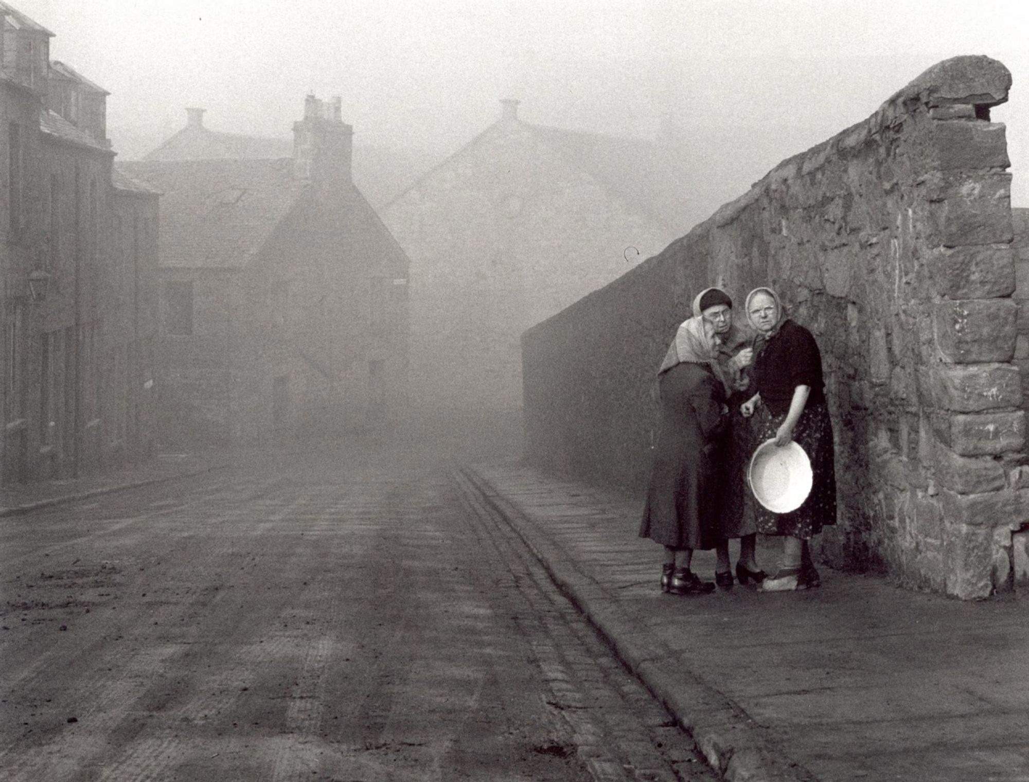 photo
Street scene, a view down a empty and foggy street, to the right, next to a wall are a group of women. They appear to be talking to each other One seems to be scowling at the photographer, she is holding a bowl.