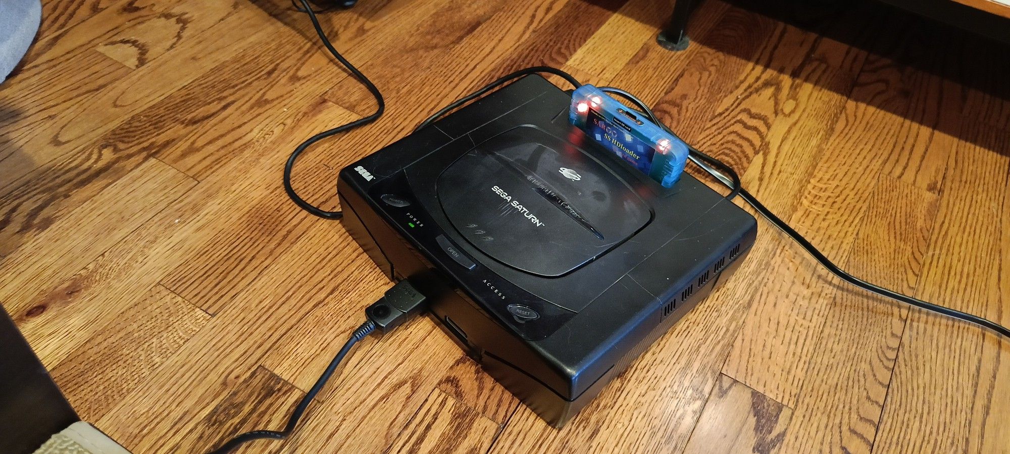 Picture of a Sega Saturn on the floor, a blue cartridge sticking from the ram expansion port