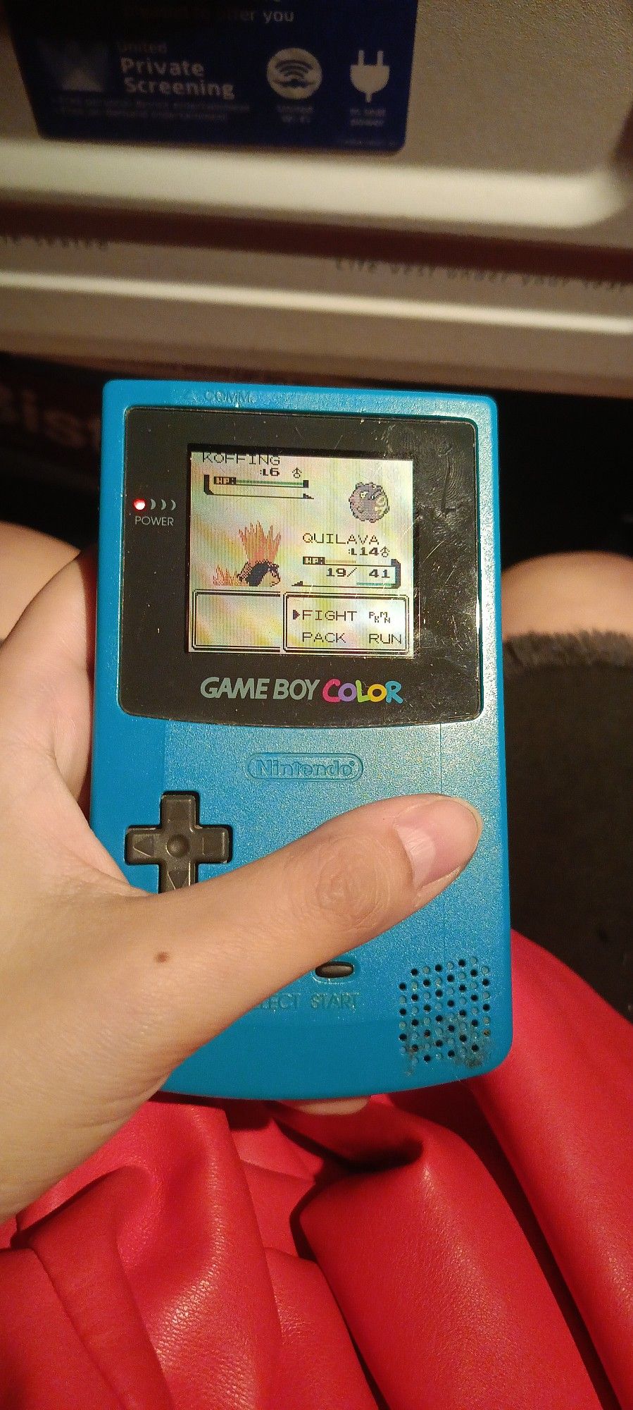 Picture of a hand holding a blue Gameboy color displaying pokemon crystal. It is very well lit.