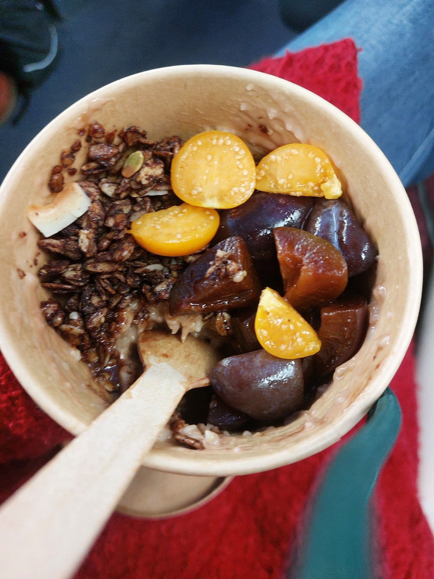 Blick in den Becher mit Porridge. Links ist im Becher der Schokocrunch, in der Mitte Physalis und rechts die Zimtpflaumen. Der Löffel aus Holz steckt schon im Porridge unterhalb der Physalis