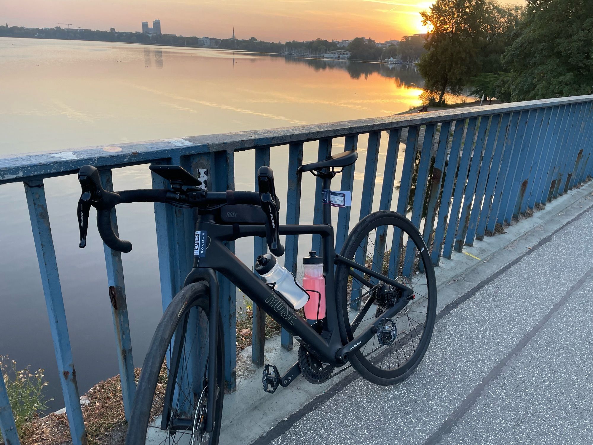 Ein Rennrad lehnt an einem Brückengeländer. Dahinter geht die Sonne auf.