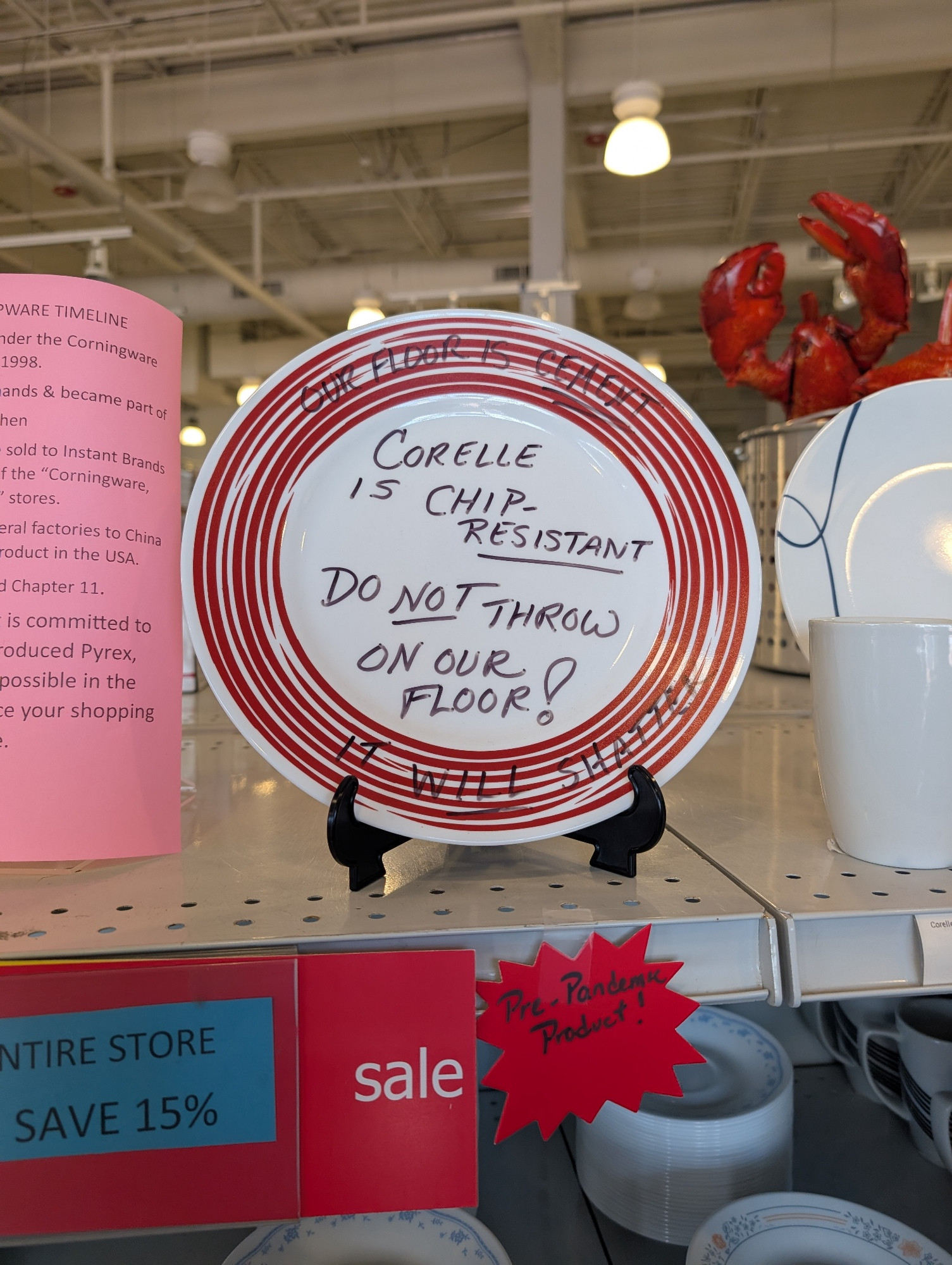 A white Corelle plate with a striped red rim sits on a retail shelf. Written in grease pencil on the plate: "Corelle is chip-RESISTANT - Do NOT throw on our floor!" And on the rim, "Our floor is CEMENT - It will shatter"