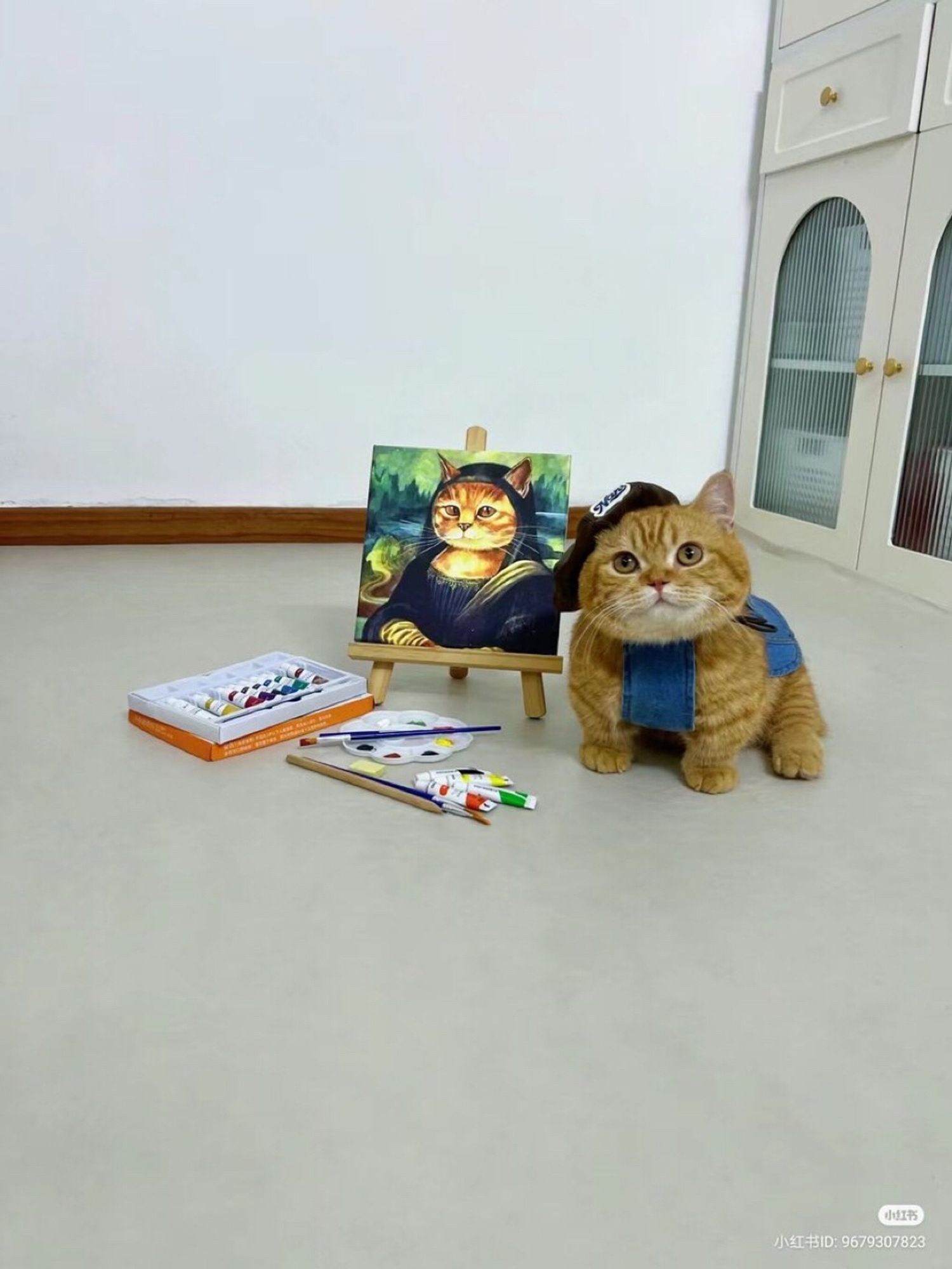 Um gatinho laranja todo fofinho usando um chapeuzinho e um cachecol, ao lado dele tem um quadro com a pintura da Mona Lisa só que com a aparência de um gatinho laranja, no chão tem objetos usados na pintura como pincéis e tintas.