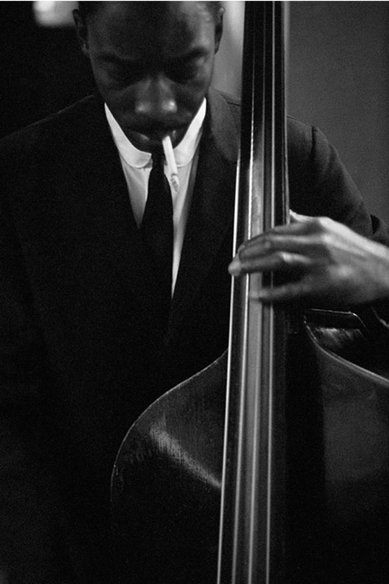 Jones, in a suit, white shirt & skinny black tie, looks down as he plays the bass. He has a cigarette in his mouth.