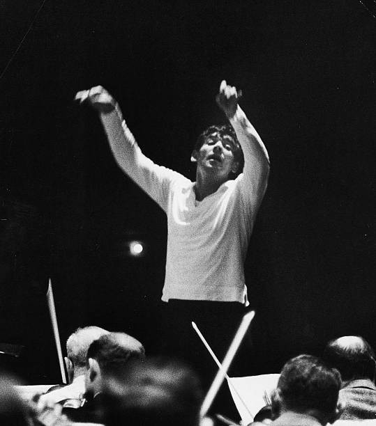 Lenny rehearses the orchestra, wearing a white shirt. His arms are high in the air.