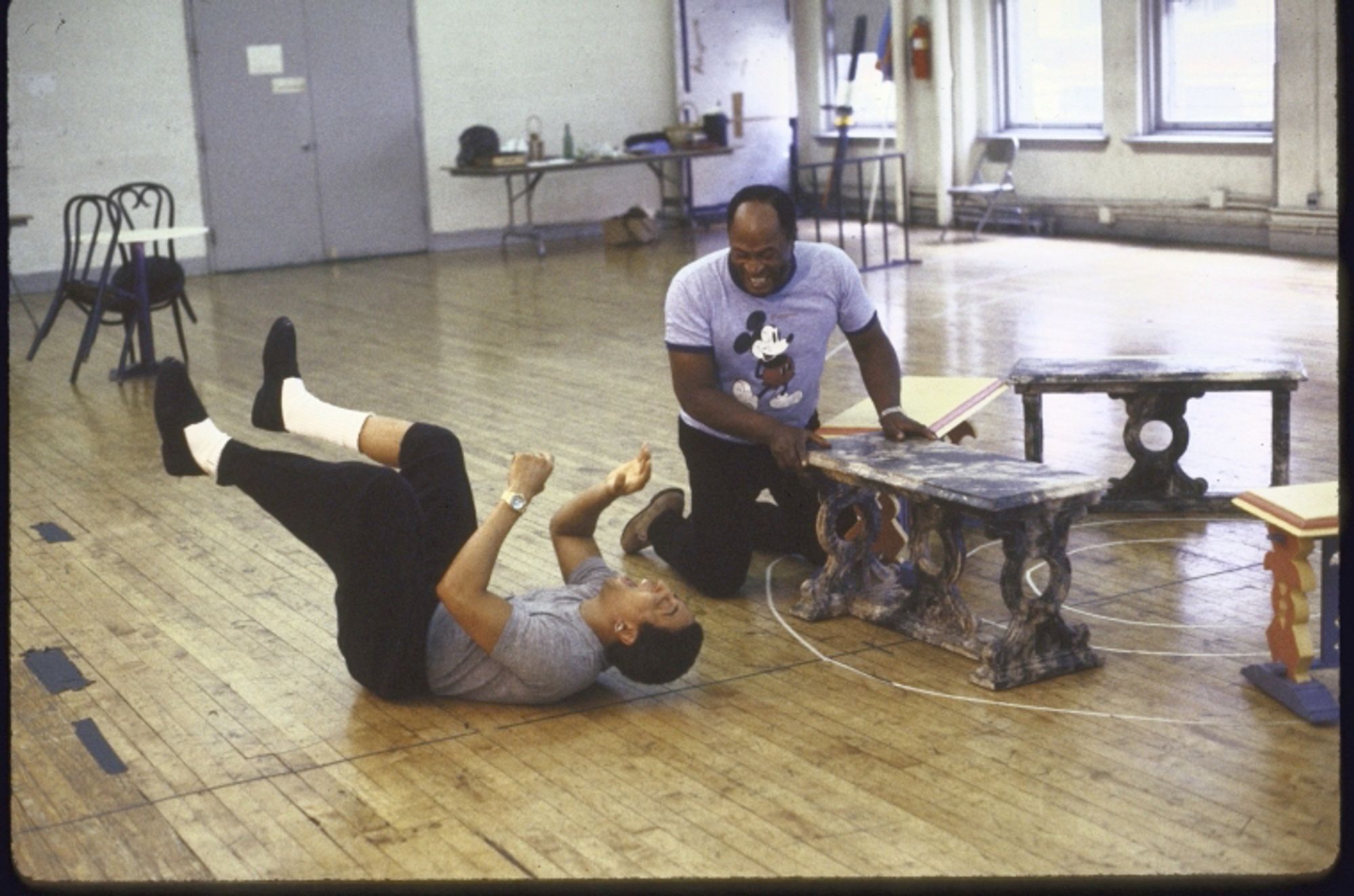 Hines & Amos in a large rehearsal space. Hines, on his back with his arms & legs in the air, is breaking up Amos, who wears a Mickey Mouse t-shirt.