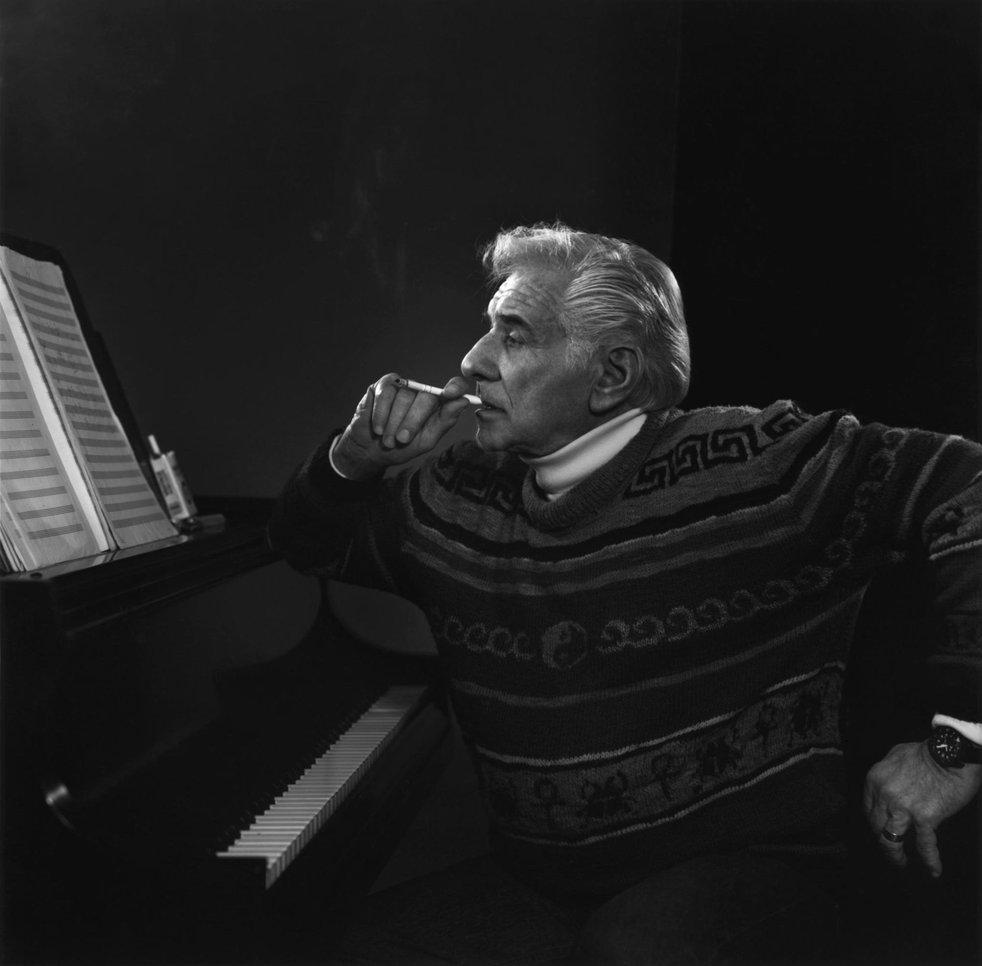 Leonard Bernstein sits at the piano, looking at a score with a cigarette in his hand. He’s wearing an impressively abstract-designed sweater.