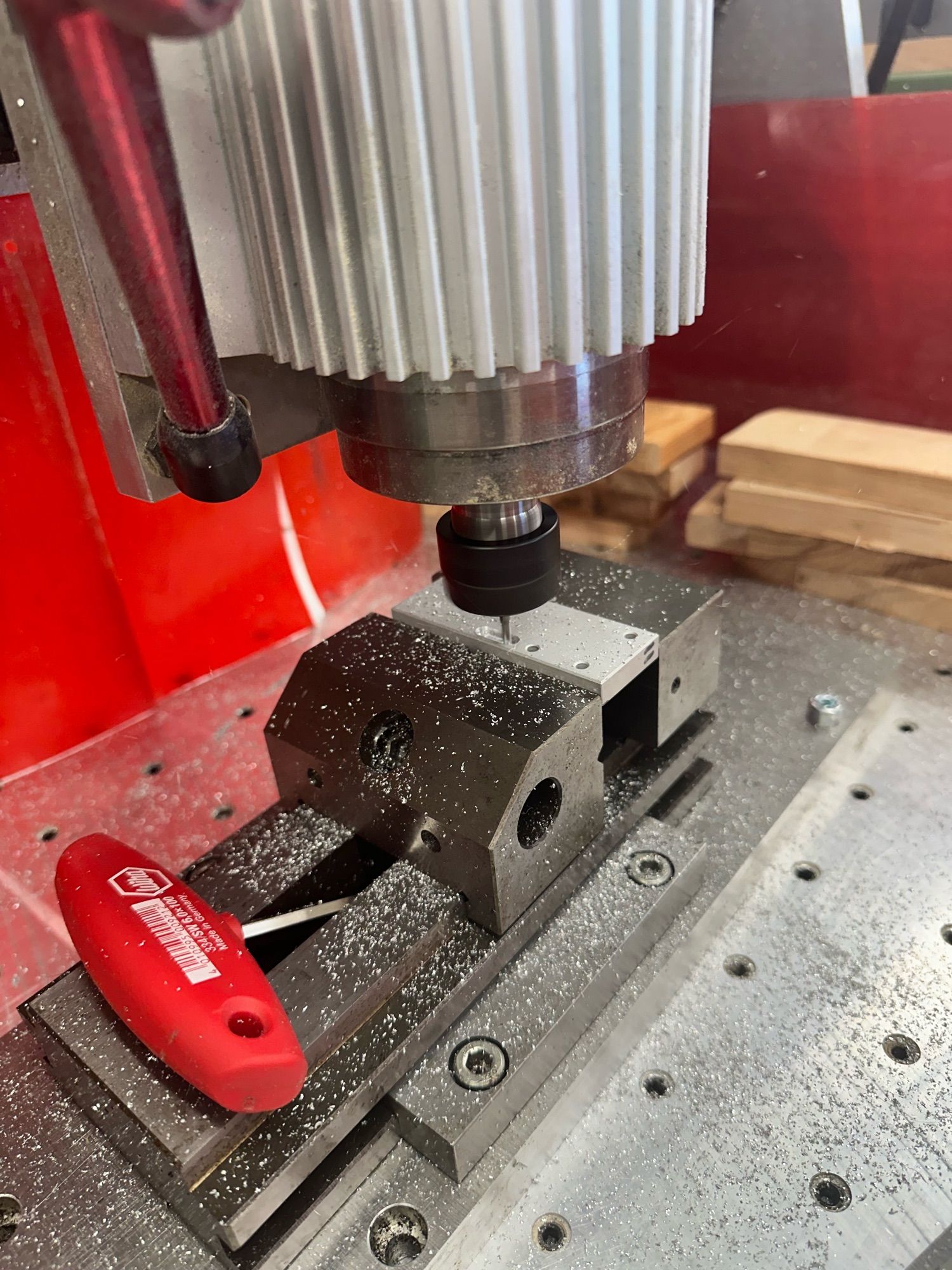 A small piece of aluminum is being held in a steel vise. There is a red handled wrench next to the vise for tightening. A milling spindle is currently cutting a countersunk slot in the piece of aluminium.