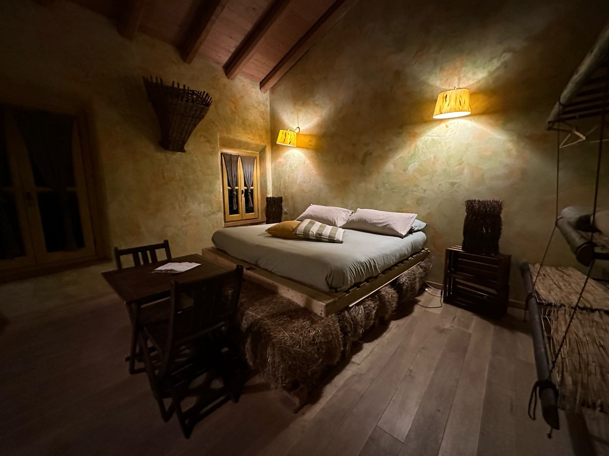 A bed in a rustikal room with e posed wooden eves and a wooden floor. The bed is sitting on several hay bales.