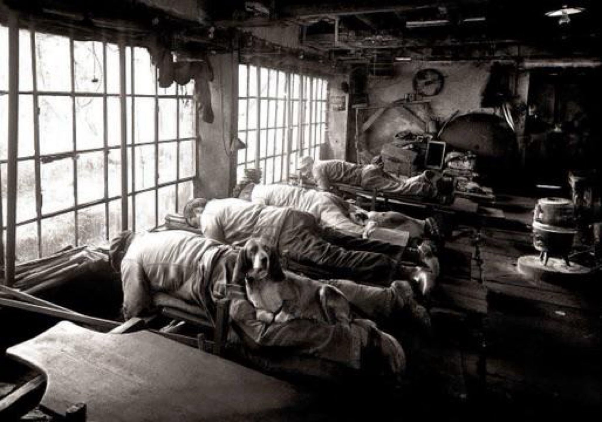 Black and white photo of four people laying on their stomach. Below them is a long shaft with grinding wheels on it and they are sharpening knives in production. Laying on the back of their legs are several dogs, ostensibly to keep them warm.