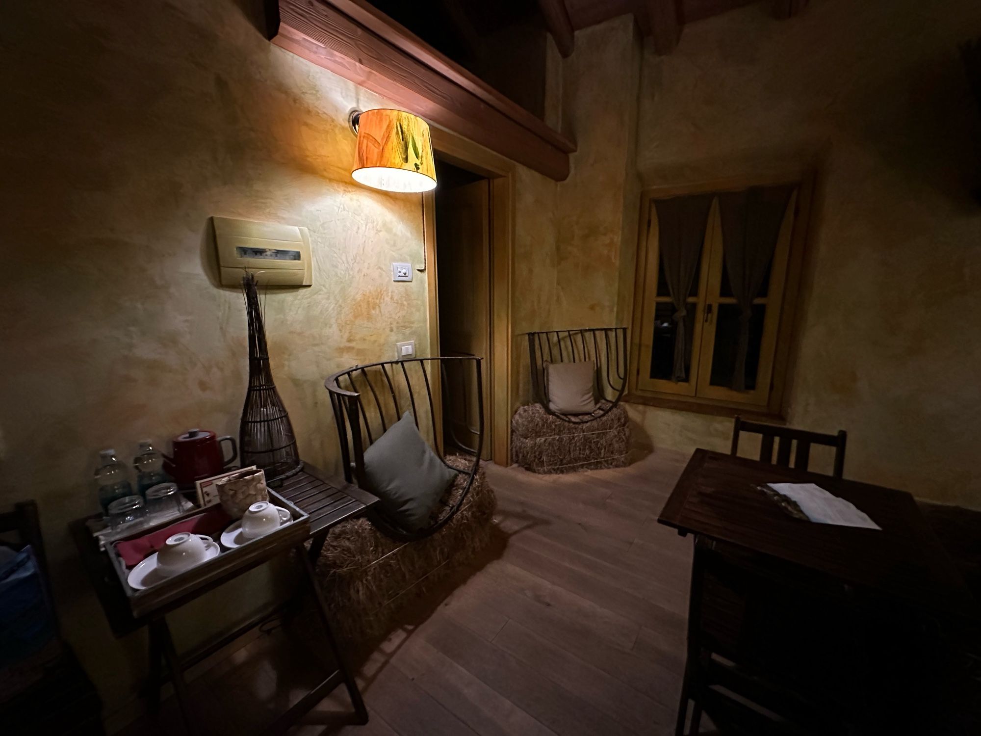 The same rustikal room, but now the other side where two chairs made from an iron grating, a pillow, and a hay bale are sitting. In the foreground is a coffee set.