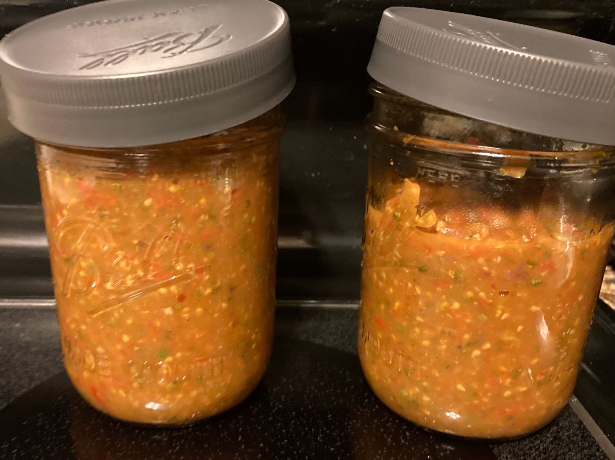 Two mostly-full jars of fresh salsa on a black glass stovetop, having just finished cooking.