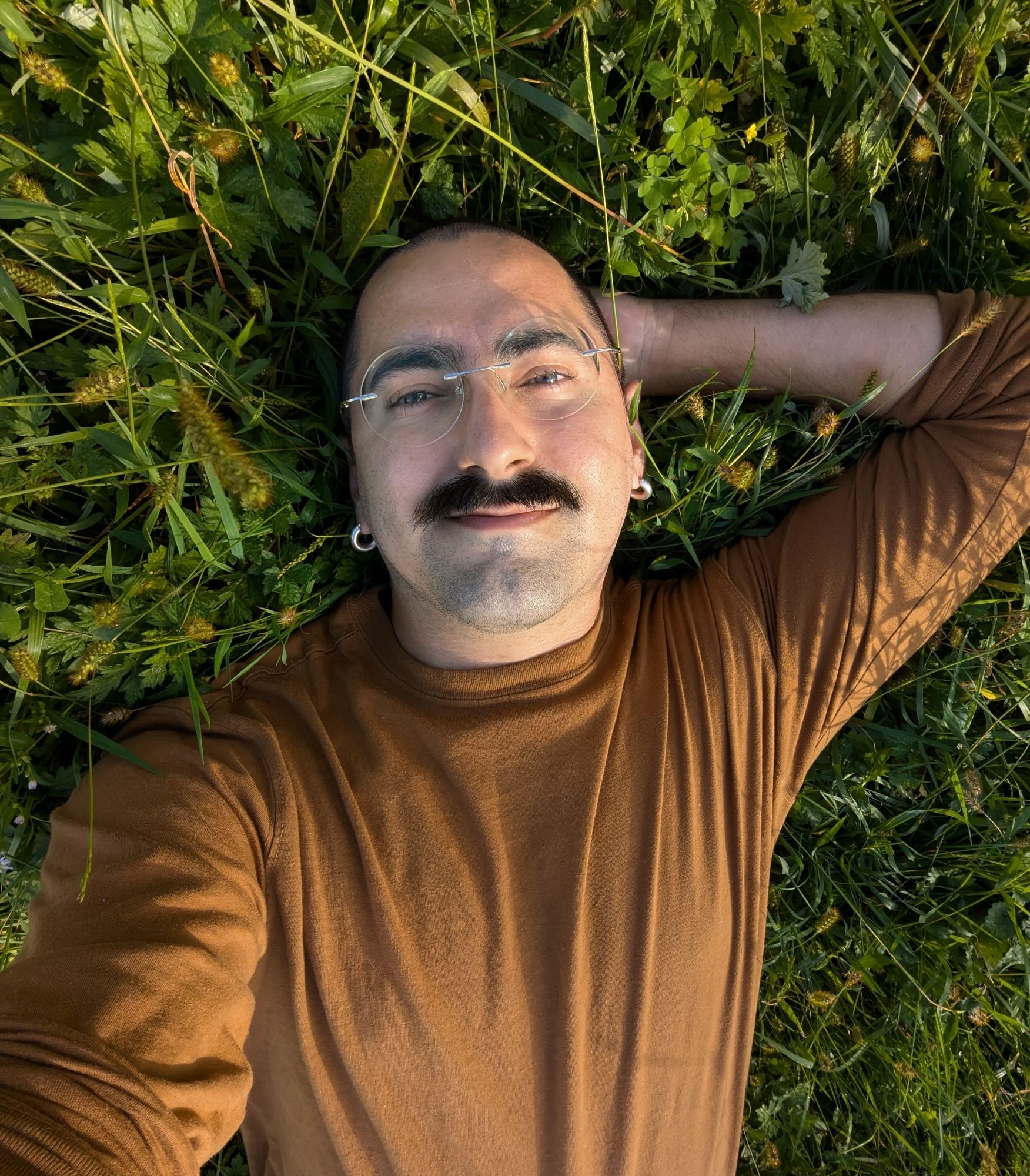 a selfie of andrei, a white person with hair buzzed short and a thick moustache, smiling and laying down in the grasses and weeds with their hand behind their head. they are wearing a long sleeved orange shirt, silver hoop earrings and round glasses. the sun is shining warm and soft in the photo, casting a golden glow.