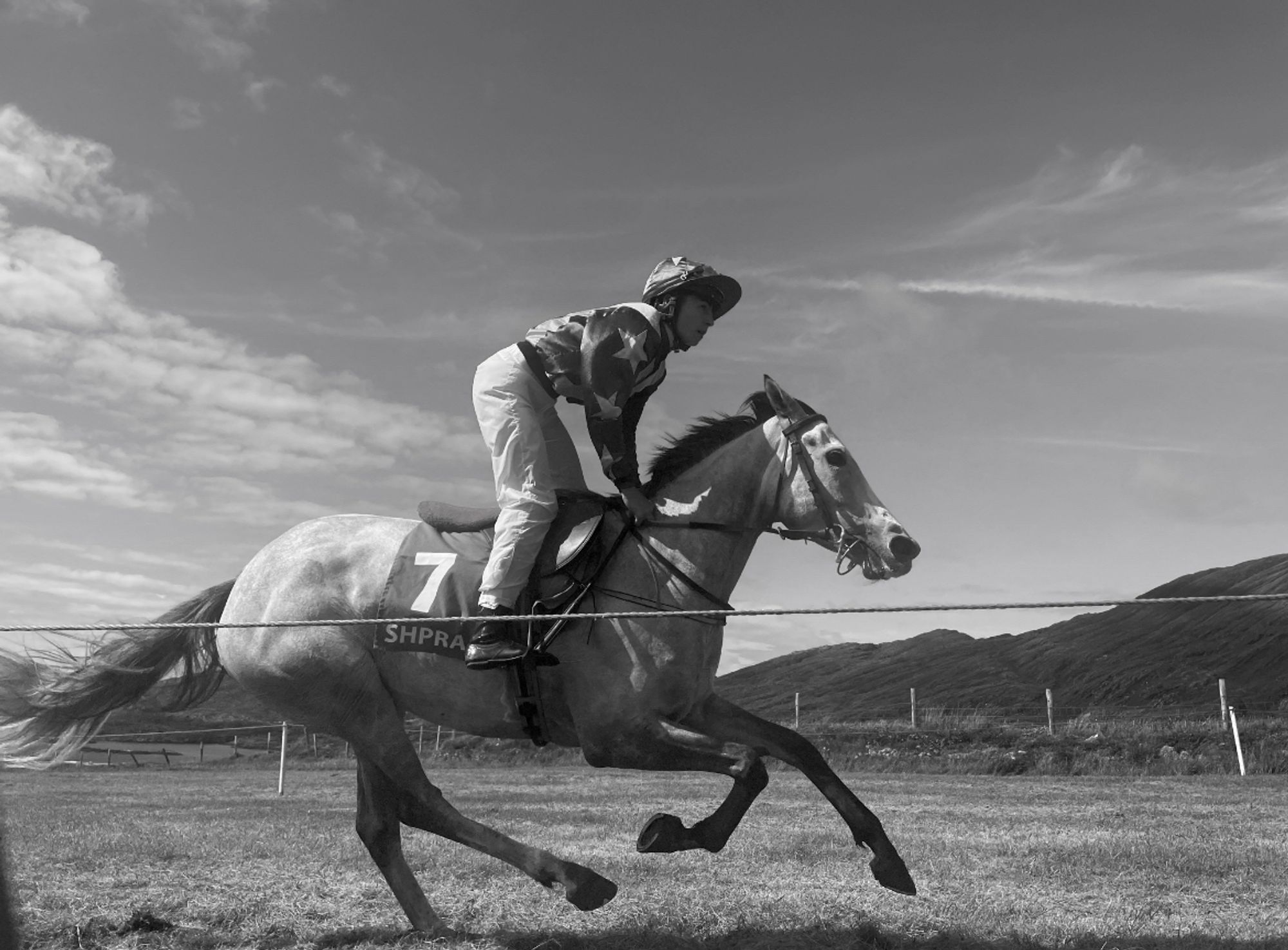 Black and white action photograph of horse and jockey racing. Sunny Summer day, with sky and hills in background.