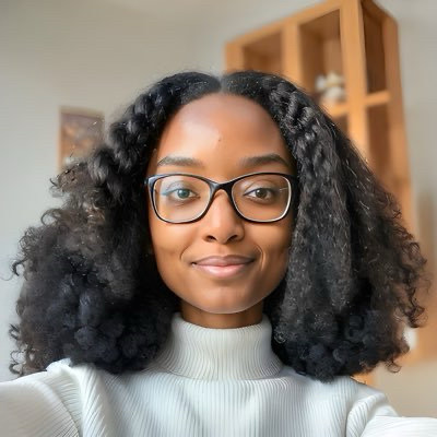 A picture of Wilough (she/her). She is wearing a white turtleneck shirt, black rimmed glasses, and has her natural hair down in a long bob. 