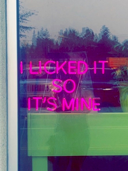 a picture i took standing in front of a shop window: 
there’s a lime green table with a vase on it in the shop.
a fuchsia neon sign hangs on the window. it says : I LICKED IT SO IT’S MINE 
you can see part of my reflection in the window