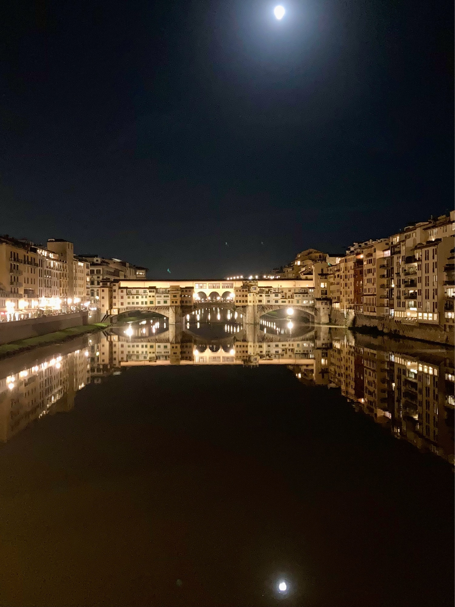 Die Ponte Vecchio in Florenz bei Vollmond. Der Nachthimmel und der Arno sind schwarz bzw dunkelbraun und der Mond spiegelt sich im Wasser