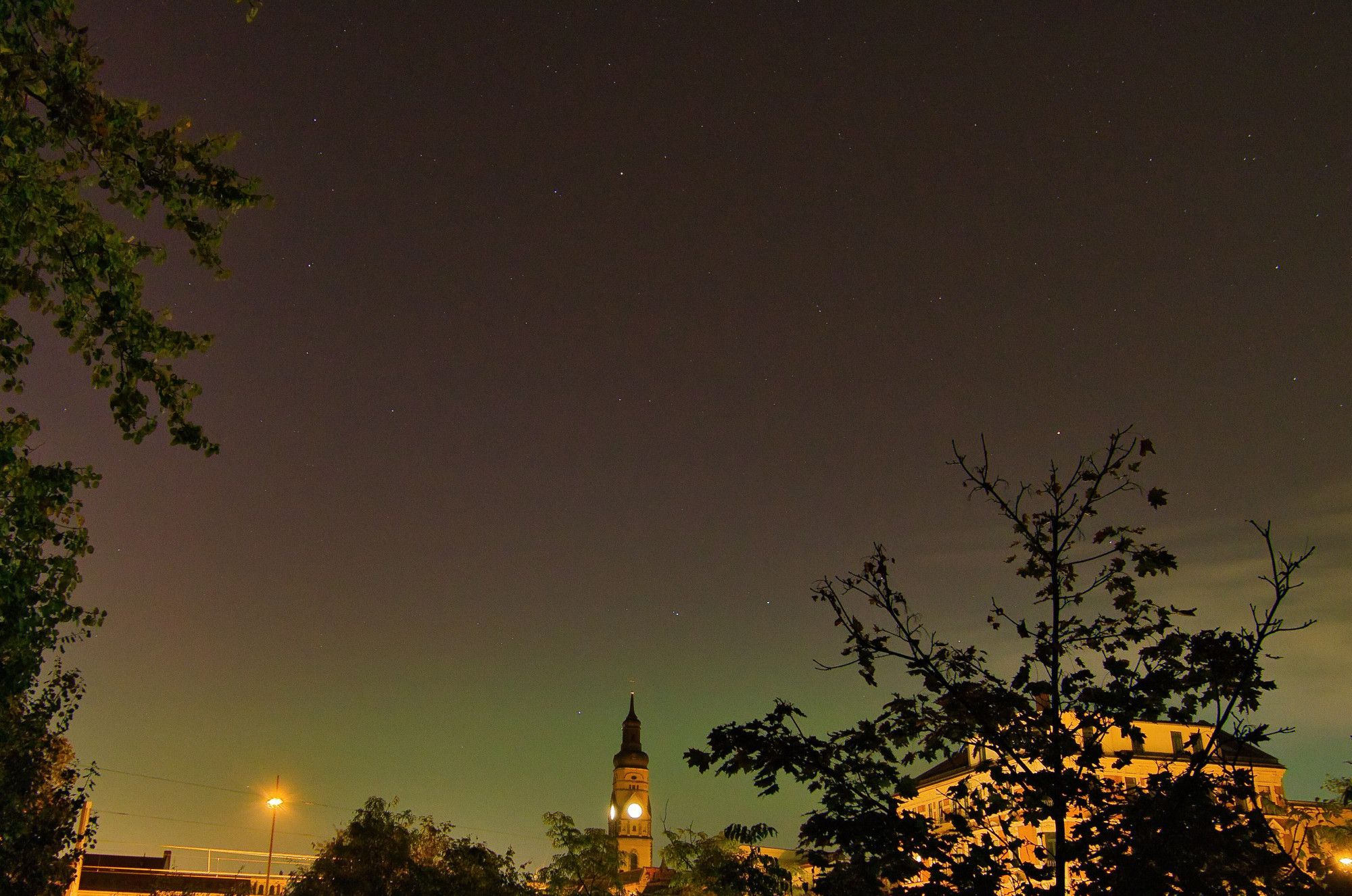 Das Bild zeigt eine nächtliche Stadtszene mit einem leicht beleuchteten Kirchturm oder einer Uhrturmstruktur im Hintergrund, umgeben von Gebäuden und Bäumen. Der Himmel ist dunkel und klar, und einige Sterne sind sichtbar. Einige Blätter und Äste der Bäume sind im Vordergrund zu erkennen. Die Straßenlaternen und die schwache Beleuchtung der Gebäude verleihen dem Bild eine ruhige, nächtliche Atmosphäre. Bei den Farben des Himmels könnte es sich um Polarlichter handeln.