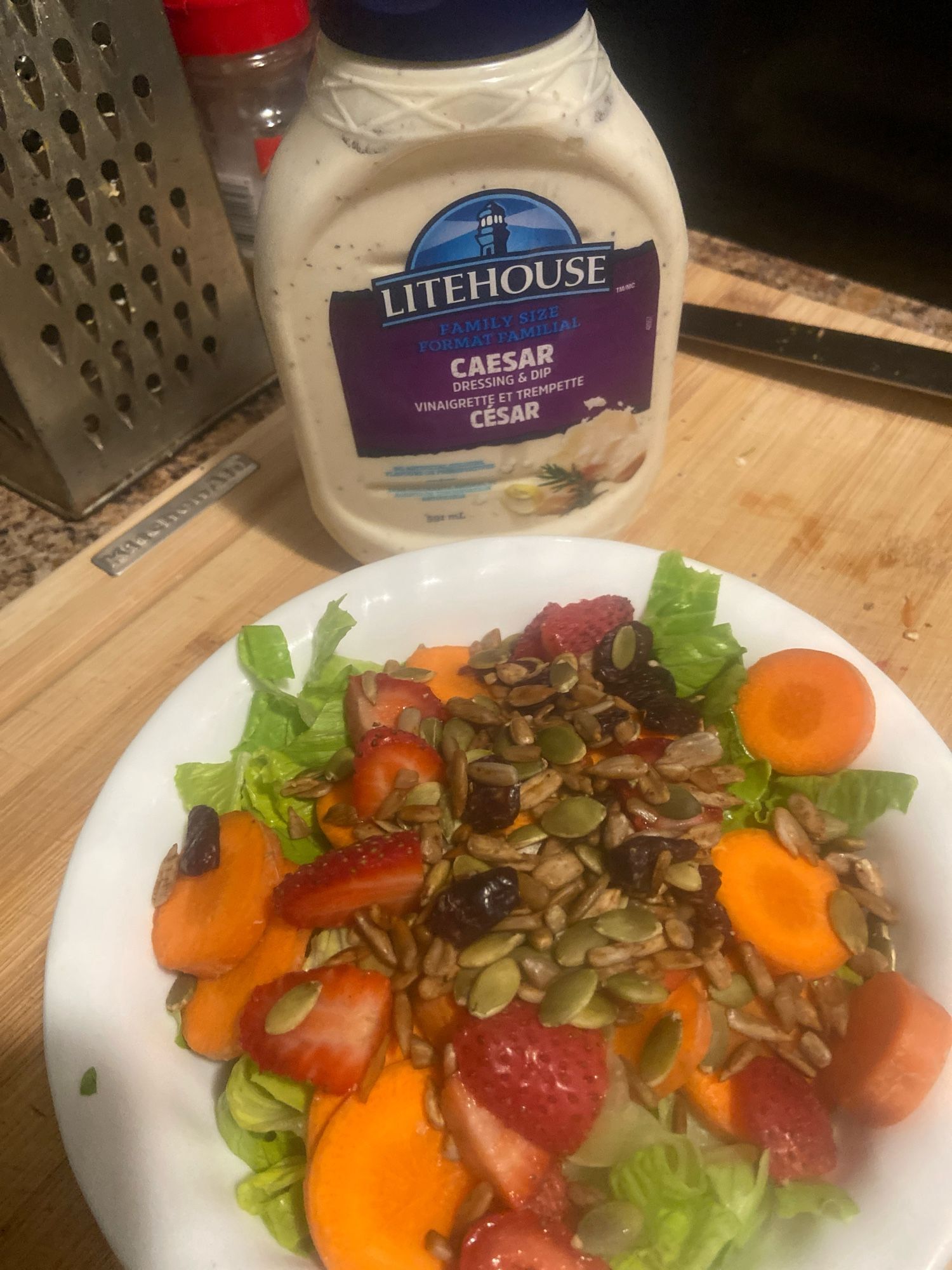 A salad with carrots, strawberries topped with nuts and cranberries right next to a bottle of Caesar dressing.