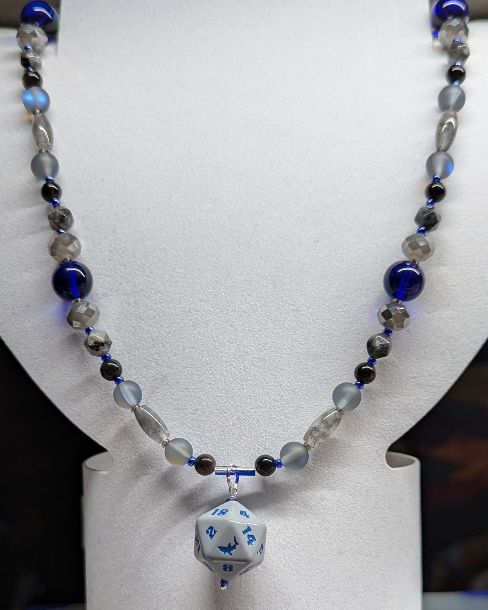 Necklace with beads in shades of gray and cobalt with a gray d20 with a shark as the 20 focal.