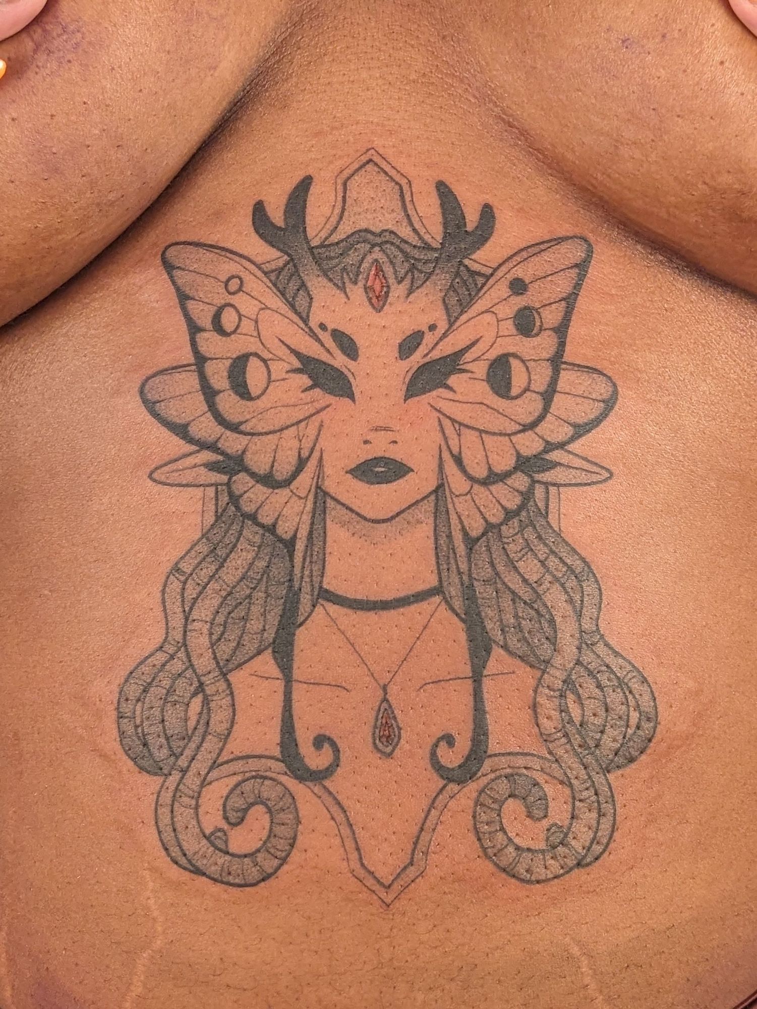 A black and grey tattoo of a neutral expressioned fae with moth wings on their face. They have small antlers and dreads.