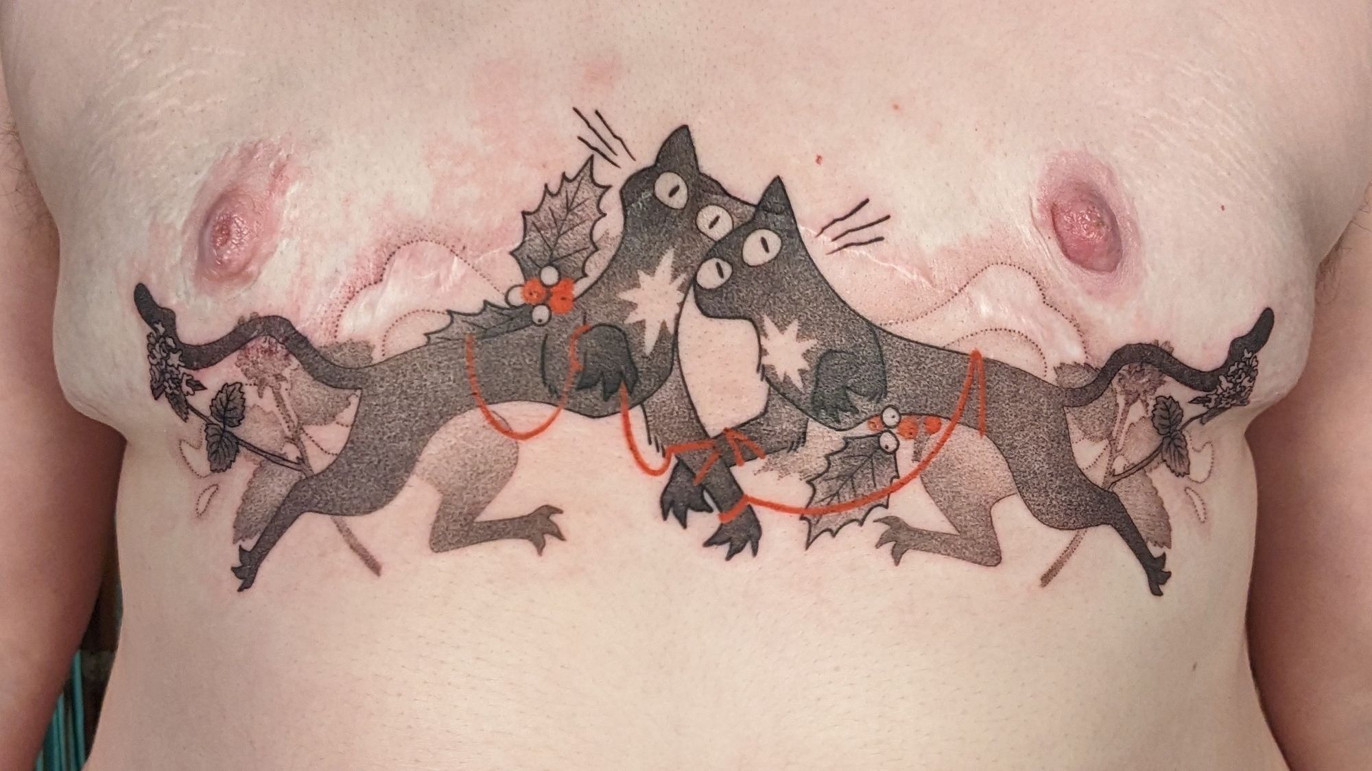 A black and grey tattoo of two cat sith tied by red strings of fate, surrounded by catnip and Holly. It is over top surgery scars