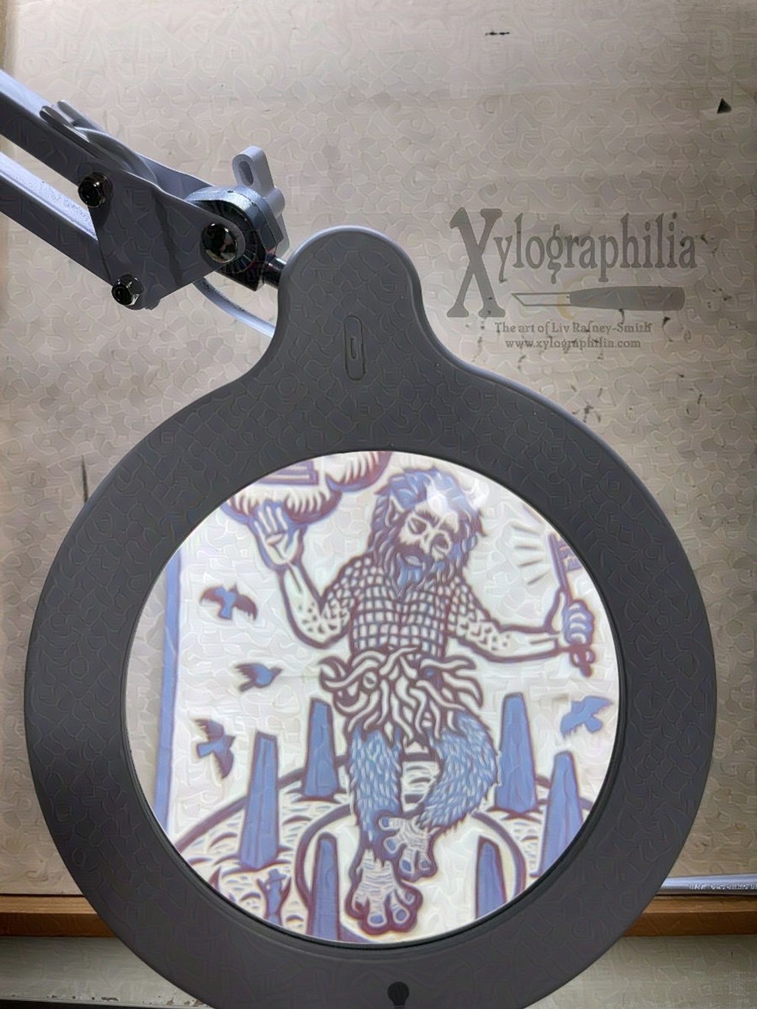 Photo looking through a magnification light at a woodcut that is almost entirely carved. Only a single eye nestled among tentacles remains to be carved. The scene is a ritual in a circle of standing stones. The primary figure a giant with large animal feet, furry legs, tentacles all over his pelvic region, and a scaled chest. His forearms, hands, and face are mostly human.