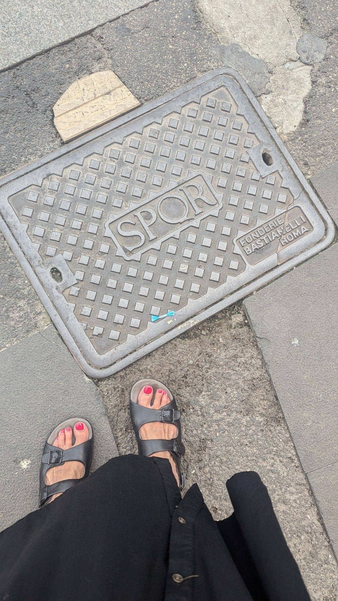 A steel utility cover embossed with the letters SPQR and the name of its foundry, embedded in a street. We see a white woman's feet in black sandals standing next to it.