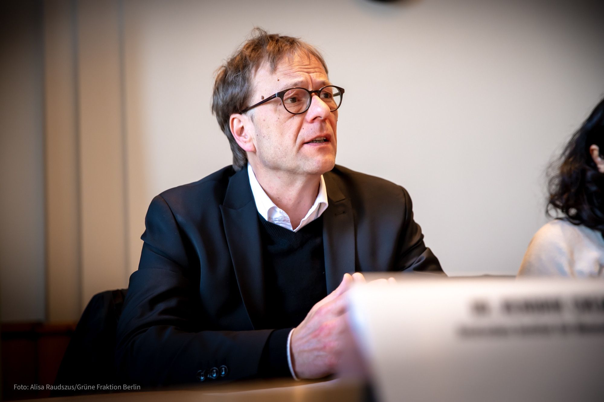 Dr. Hendrik Cremer sitzt an einem Tisch und spricht mit ernstem Gesichtsausdruck. Er trägt Brille und einen schwarzen Pullover mit schwarzem Sakko.