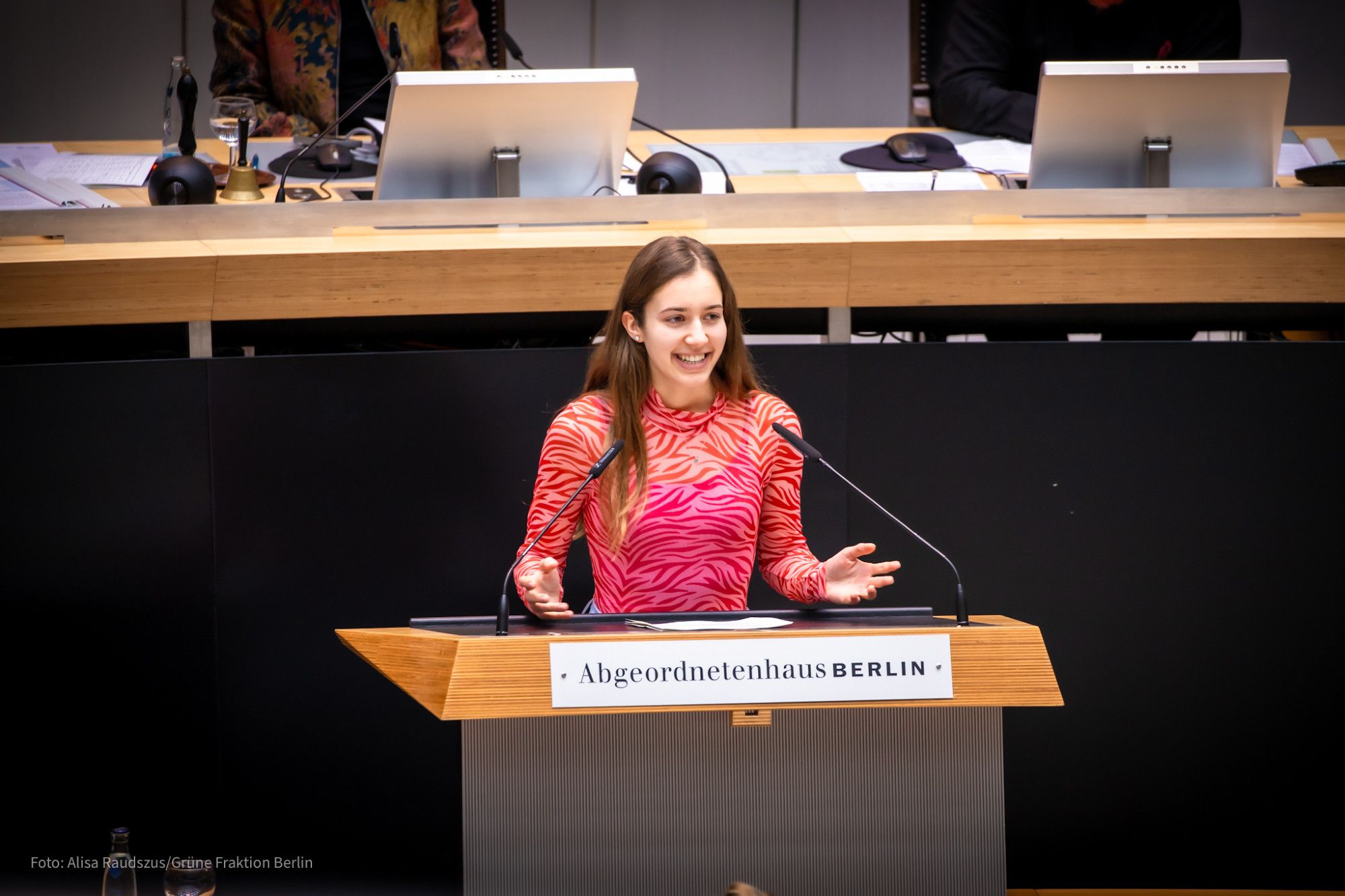 Klara Schedlich steht am Redepult des Abgeordnetenhauses und hält eine Rede. Sie trägt ein pinkes Langarmshirt, ist sehr jung und lacht herzlich.