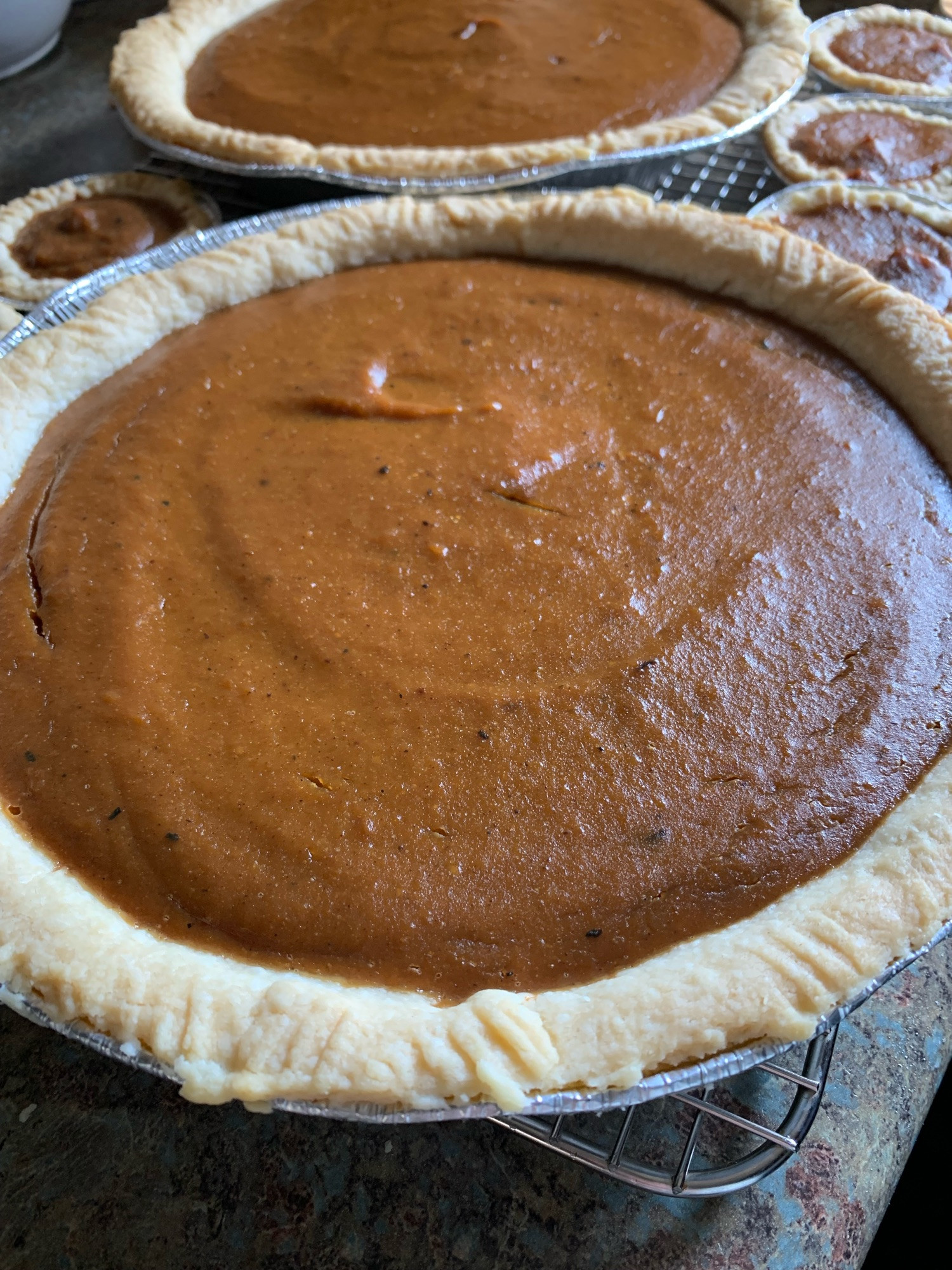 A close up of a pumpkin pie, showcasing the dark orange filling, sitting on a cooling rack, with another pie and tarts in the background.