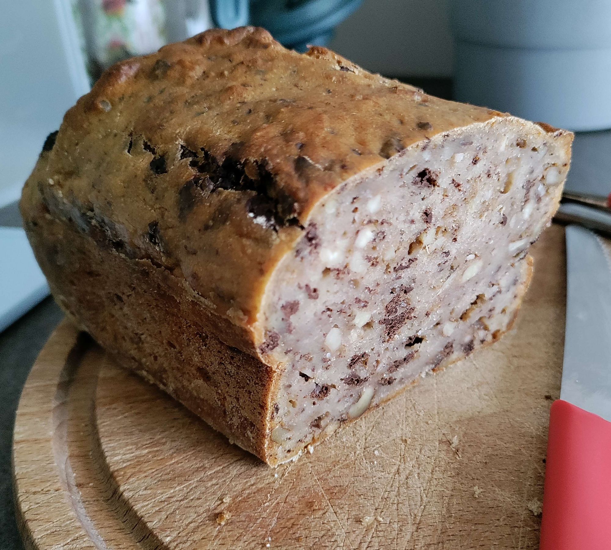 Ein Laib Bananenbrot, welches an einer Seite aufgeschnitten wurde. Rechts vom Bananenbrot ist ein Messer. Beide sind auf einem Holzbrett.