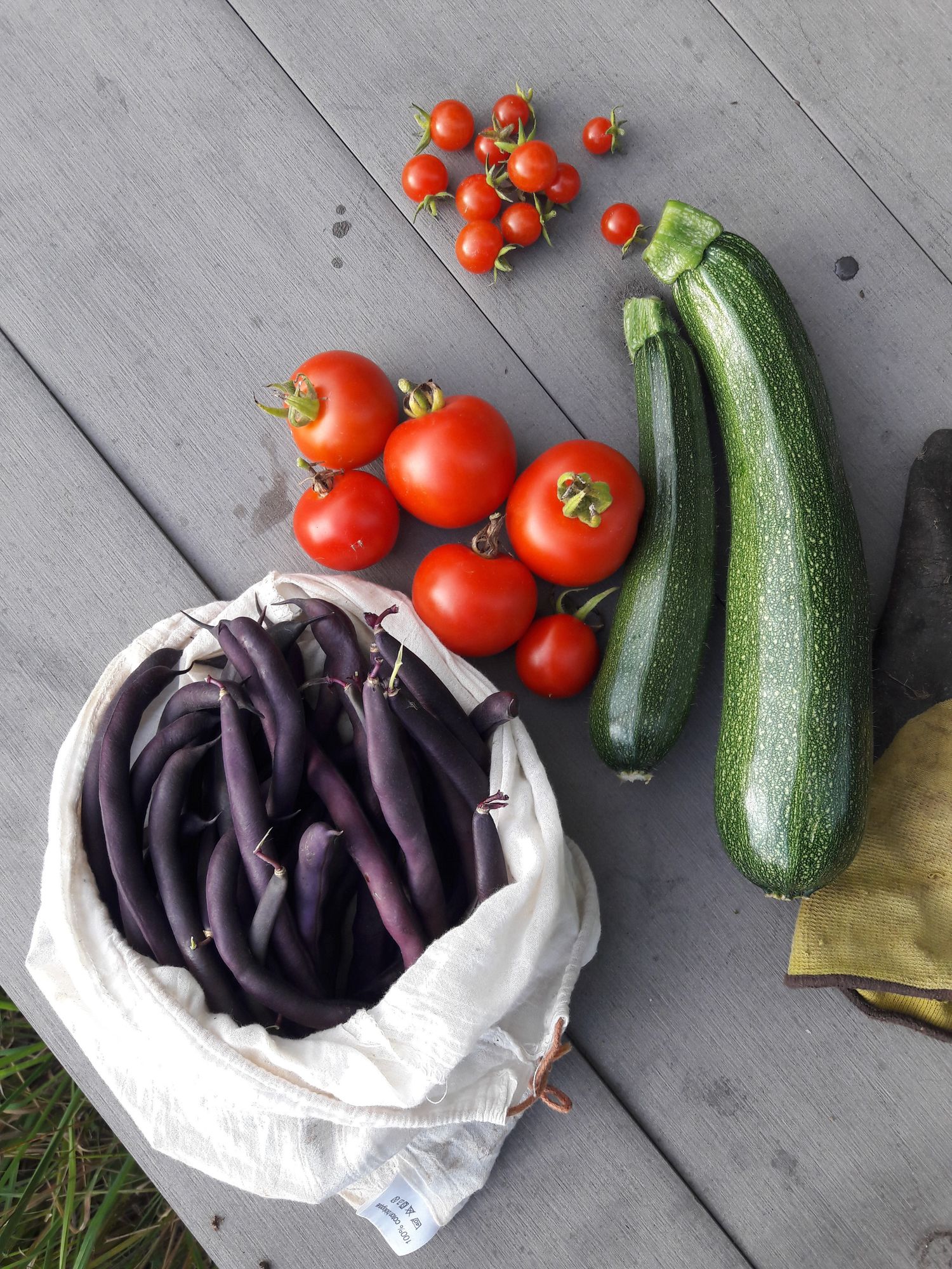 Une table de jardin prise du dessus sur laquelle sont posées des tomates, des petites tomates, deux courgettes et un sac de haricots violets