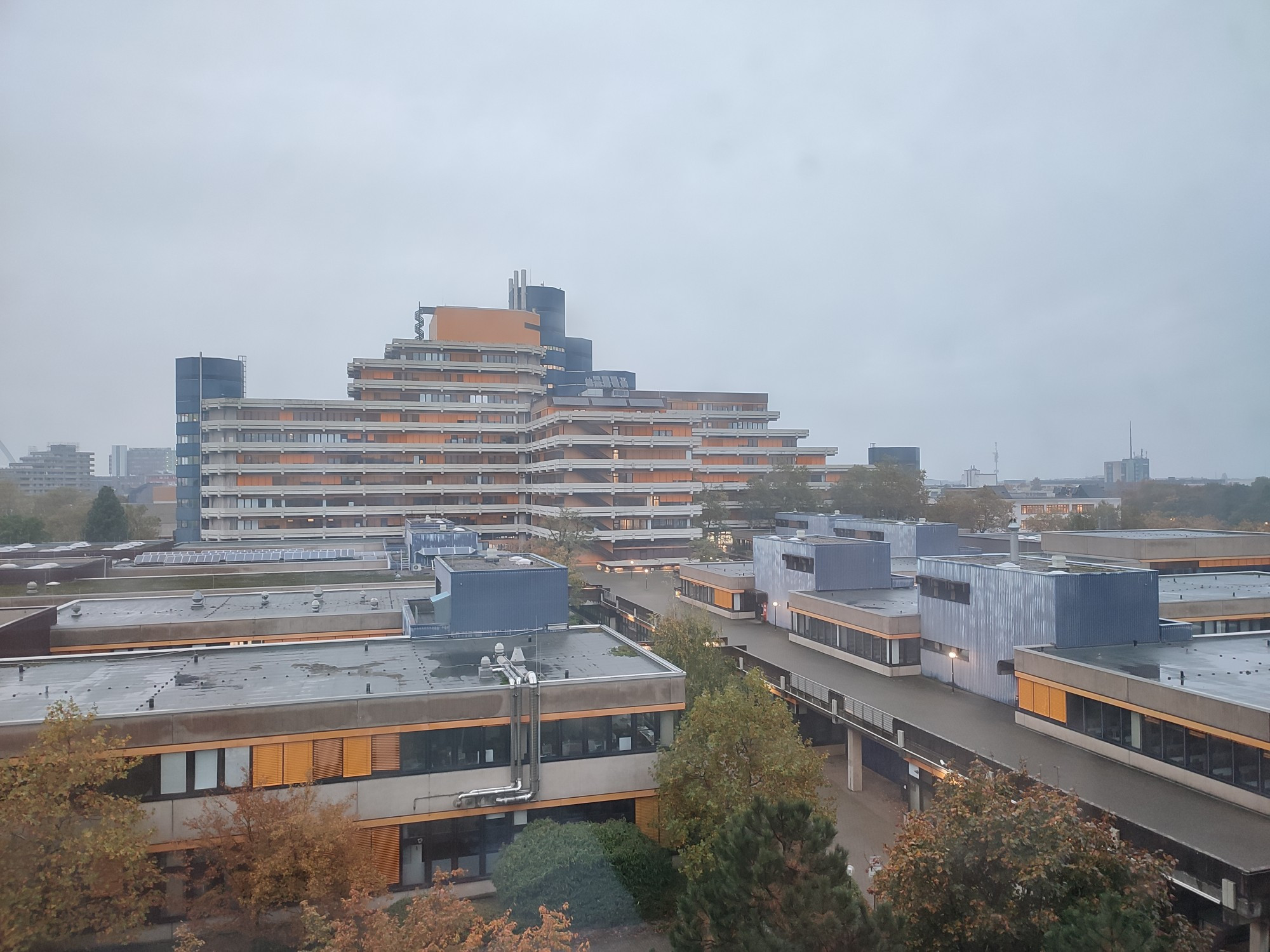 Tristes mehrstöckiges Gebäude vor tristen flachen Gebäuden, beide aus grauem Beton mit orangefarbenem Elementen, unter einem bedeckten grauen Himmel.