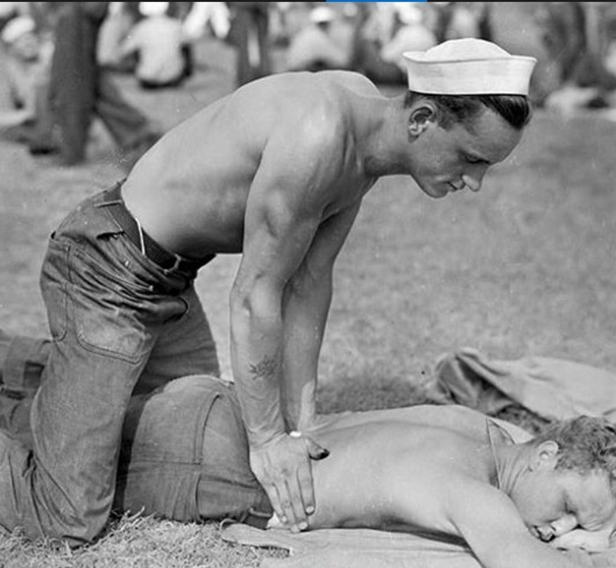 Black and white photo of a shirtless WWII era sailor in a sailor hat suggestively rubbing his fellow sailors back.