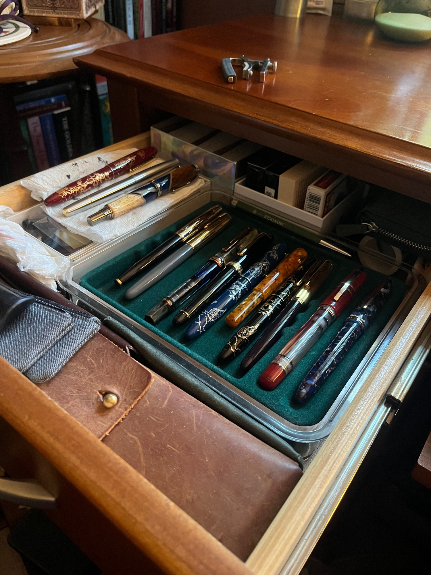 A wooden desk drawer, open to reveal a green velvet tray lined with ten fountain pens. Three more rest on an ink-stained paper towel next to the tray. The compartments of the drawer are loosely filled with single pen sleeves, more ink-stained paper towels for cleaning purposes, a pencil case, a couple of decks of playing cards, and other random bits.