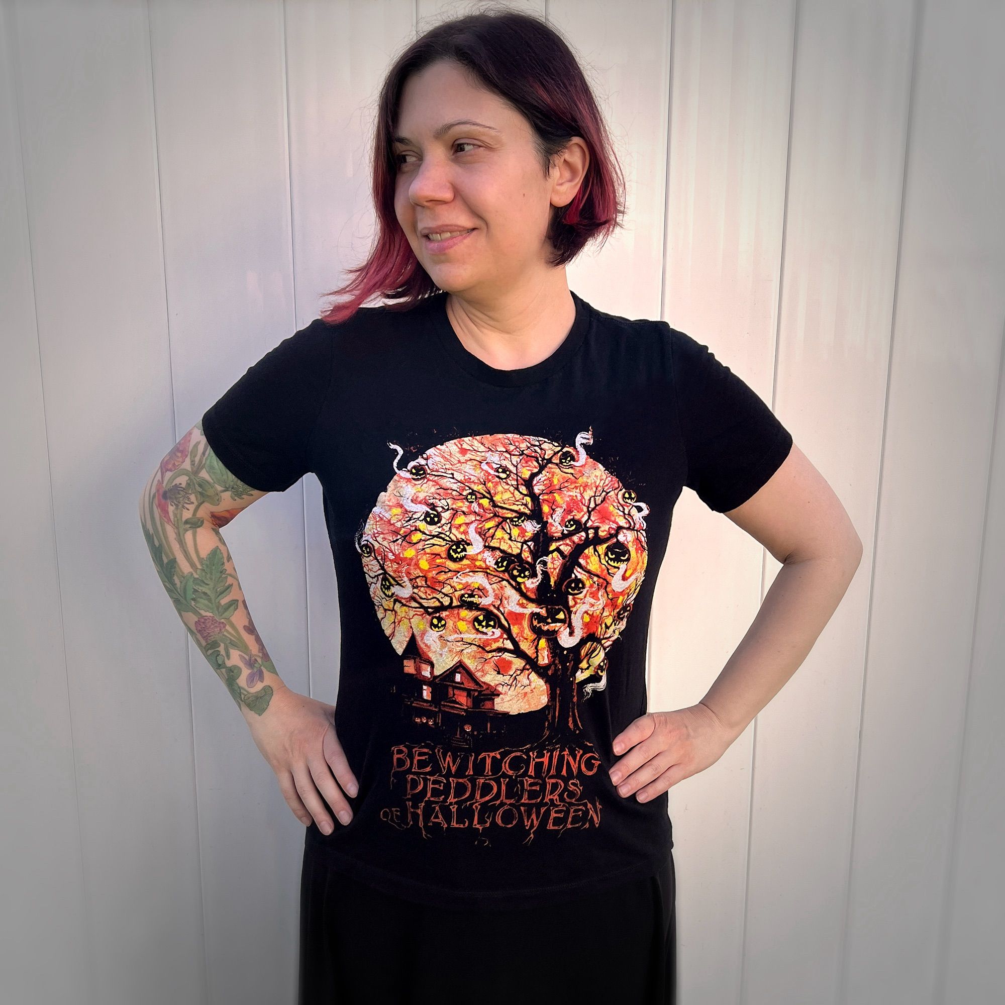 The artist modeling a screen printed t-shirt. Black shirt with art of a dark Victorian house and scraggly tree silhouetted against a big full moon. Many different jack o lanterns hang from the tree. Below it says “bewitching peddlers or Halloween” as if the letters are growing from the roots of the tree.