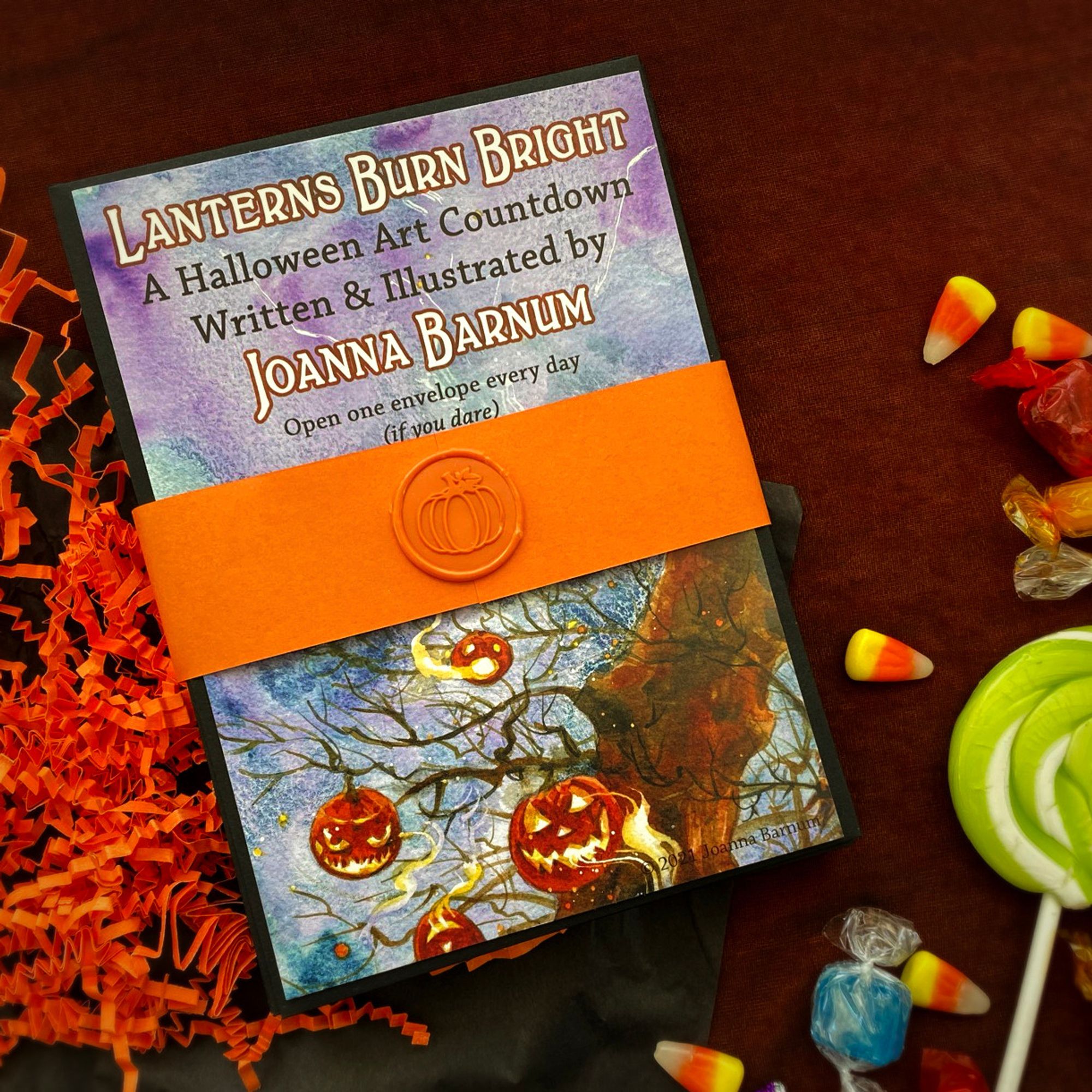 A photo of a "Halloween Art Countdown," a stack of envelopes with an illustrated cover, bundled together with a decorative band and wax seal. The cover says "Lanterns Burn Bright" with an illustration of jack-o-lanterns.