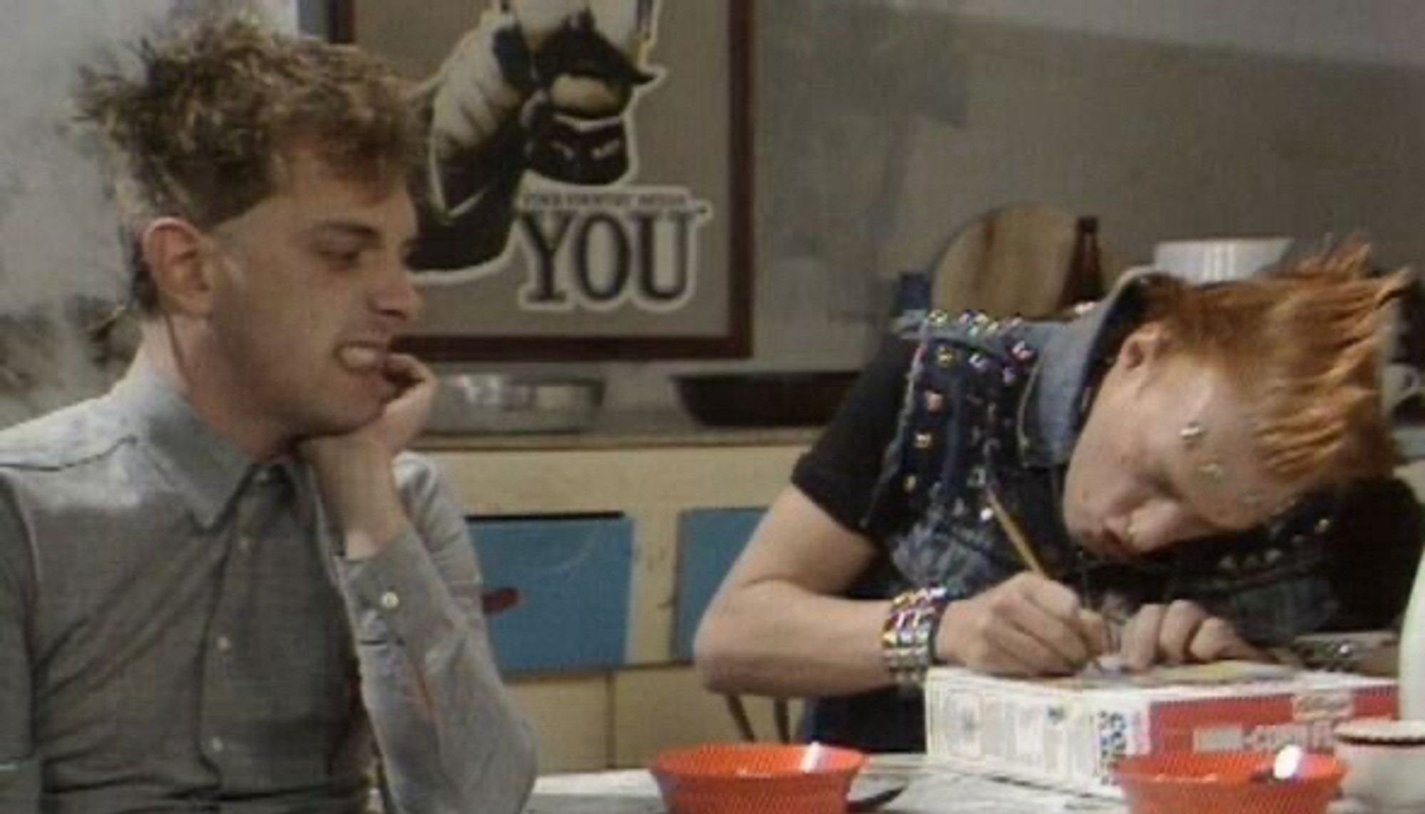 Rik and Vyvyan at the kitchen table. Vyv is hunched over a box of Corn Flakes with a pen, concentrating on writing. Rik is glaring at him with disdain.