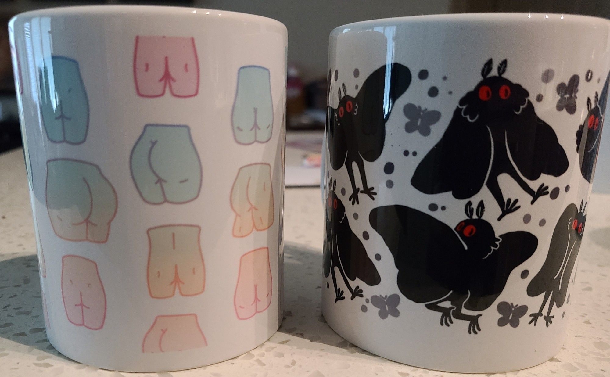 2 coffee mugs, one with pastel butts and one covered in cute little mothmen, some of which are shaking their little cryptid booties