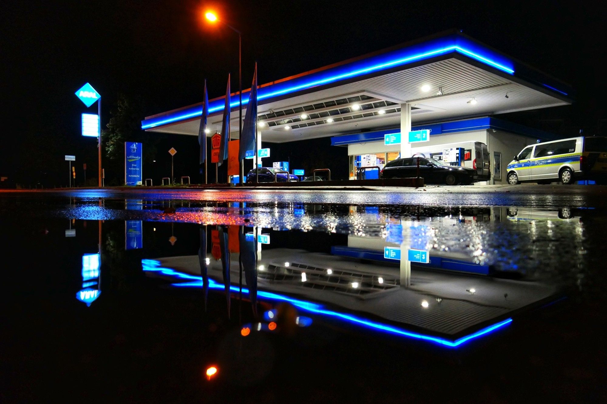 Das Bild zeigt eine Aral-Tankstelle bei Nacht. Die Tankstelle ist hell erleuchtet, hauptsächlich in Blau- und Weißtönen, was einen starken Kontrast zur dunklen Umgebung bildet. Es gibt mehrere Zapfsäulen für Fahrzeuge, an denen Autos betankt werden. Ganz rechts ist ein Polizeibus zu sehen. Die Beschilderung der Tankstelle ist ebenfalls beleuchtet und zeigt die Marke und die Preise an, obwohl der Text in dieser Ansicht nicht lesbar ist. Das Ganze spiegelt sich in einer Pfütze.