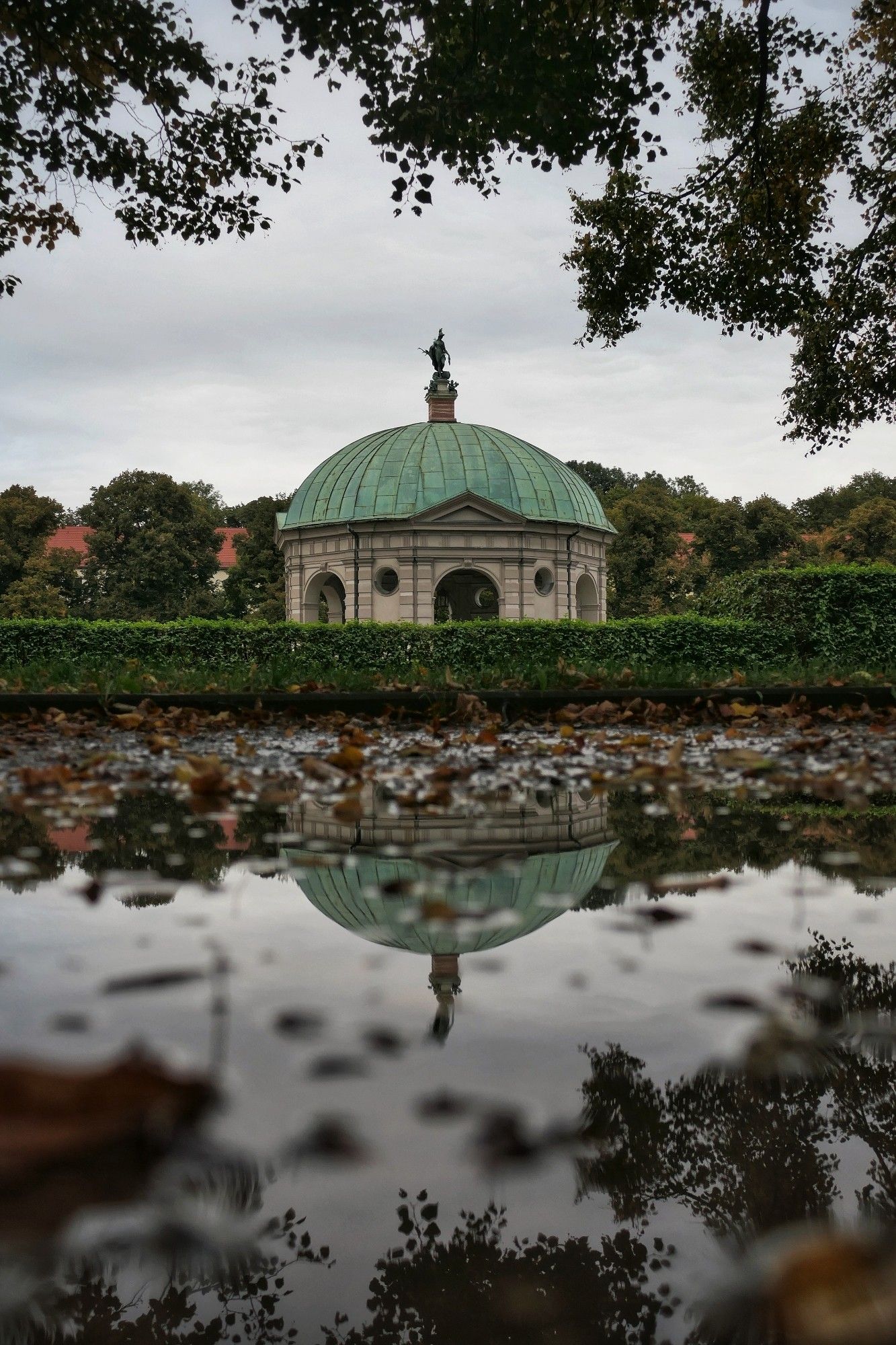 Das Bild zeigt einen Pavillon mit einer Kuppel und einer Statue auf der Spitze, umgeben von üppigem Grün in einem Park. Das Gebäude hat mehrere Bögen und scheint aus Stein zu bestehen. Die grüne Kuppel ist mit einem dekorativen Abschluss versehen. Im Vordergrund spiegelt sich das Gebäude in einer Pfütze. Der Himmel ist bewölkt und grau, was der Szene eine ruhige und leicht melancholische Atmosphäre verleiht. Das Bild ist durch Zweige von Laubbäumen eingerahmt.